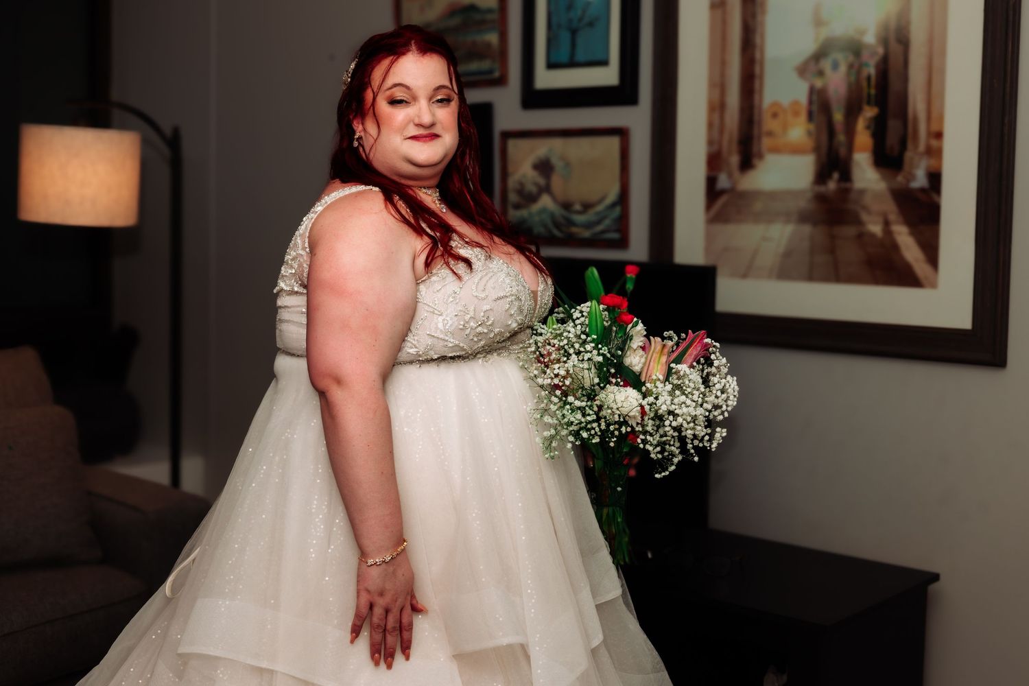 A plus-size bride with red hair in a layered white ballgown holds a bouquet indoors.