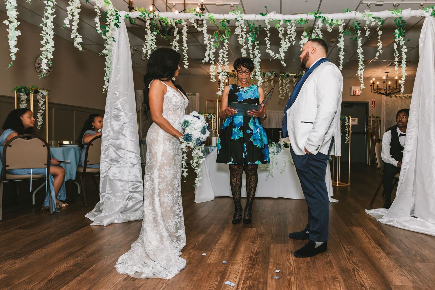 Bride and groom exchange vows during indoor wedding ceremony beneath hanging white floral greenery arch.