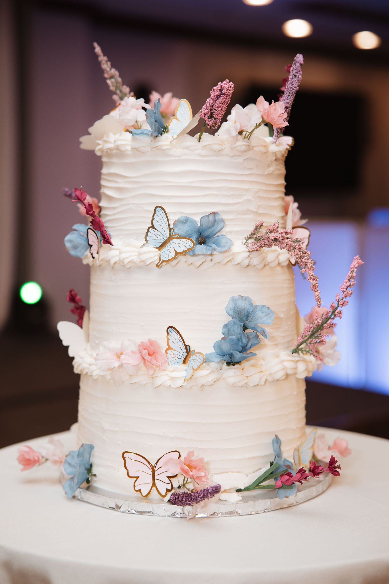 Three-tier white wedding cake with blue flowers, pink blooms, and gold butterfly decorations.