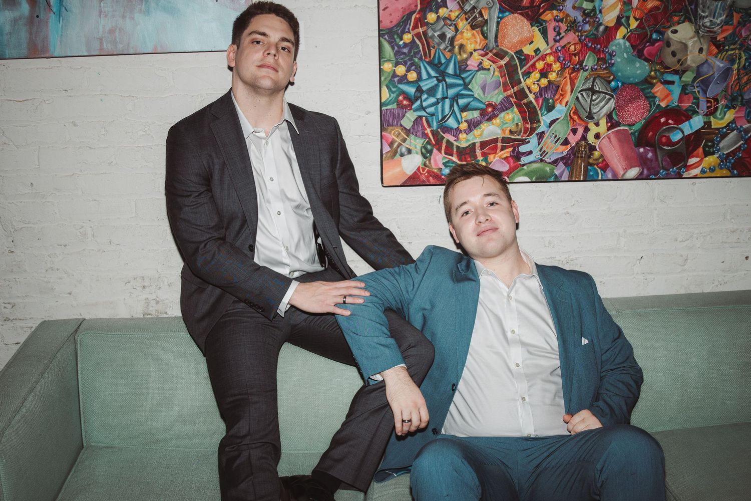 Two well-dressed young men in suits posing casually in front of colorful abstract artwork.