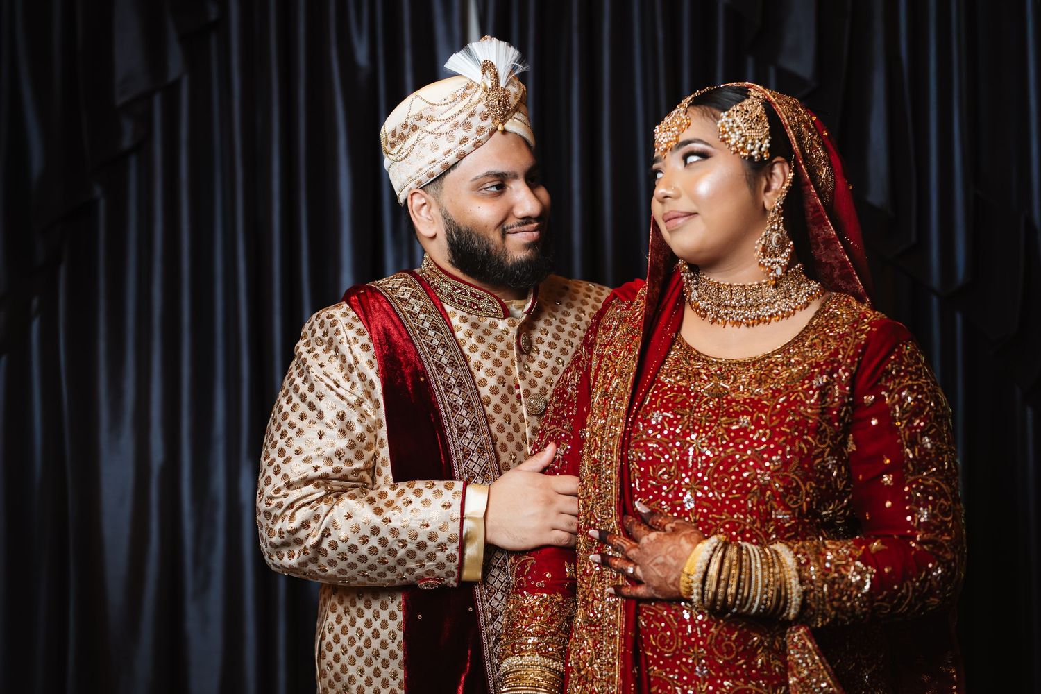 Indian wedding couple in ornate traditional bridal outfits, bride in red lehenga and groom in gold sherwani.