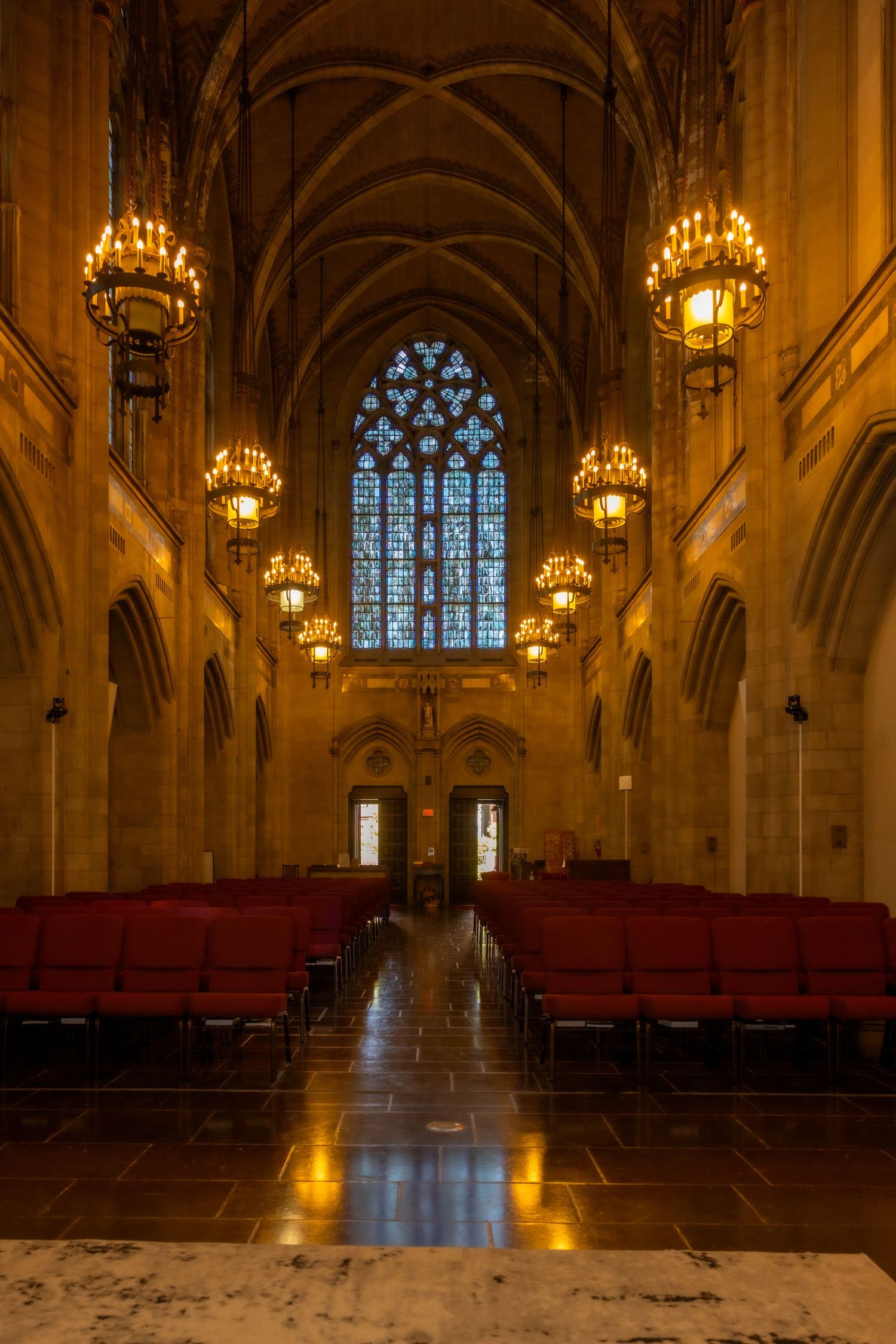 Grand Gothic cathedral nave with vaulted ceiling, glowing chandeliers, red seats, and stained glass window.