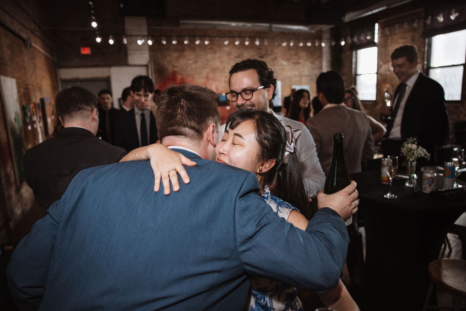 Guests share warm hugs at an indoor cocktail party in a rustic brick venue.