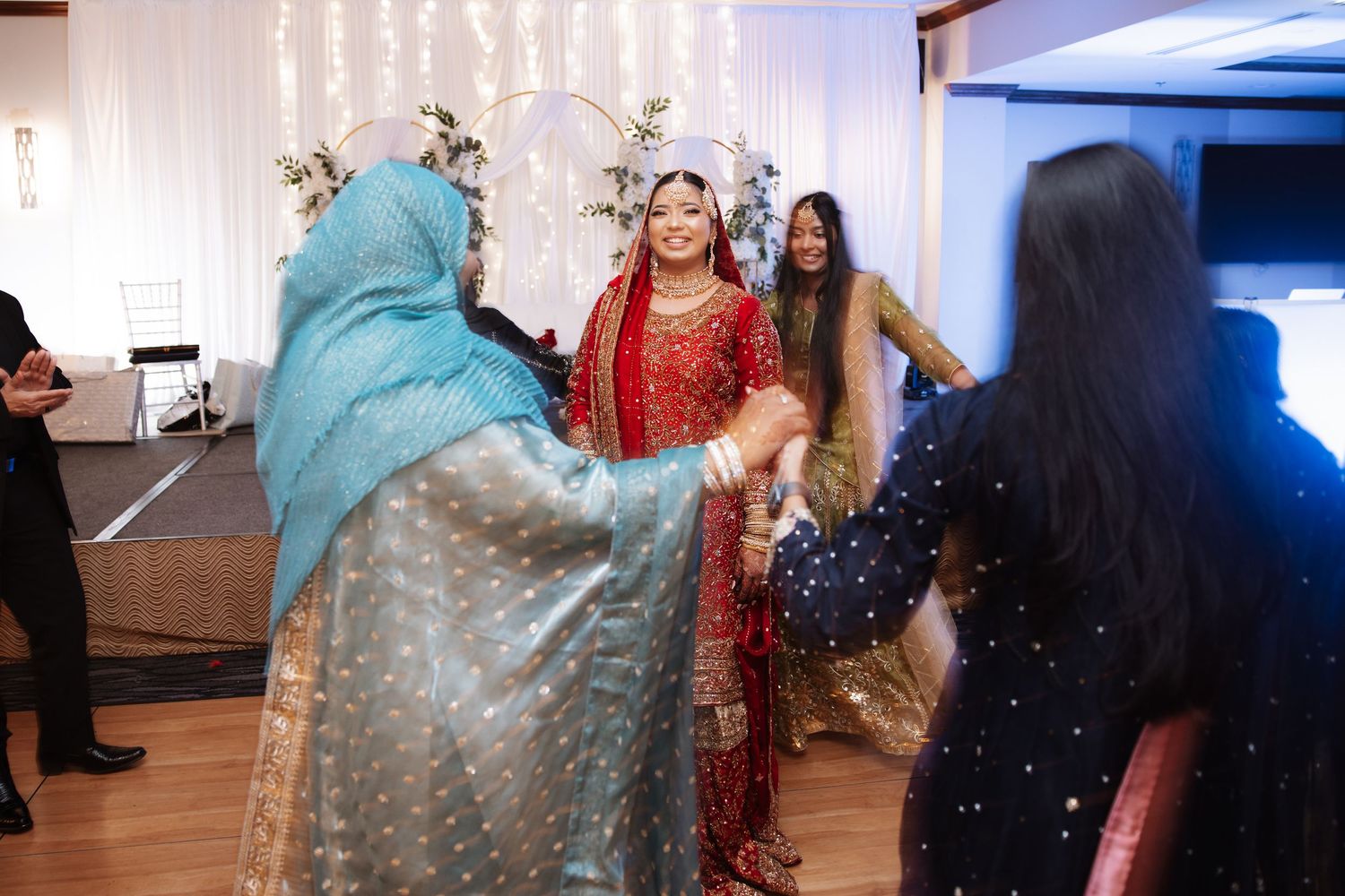 Bride in red traditional Indian bridal outfit greeting guests in colorful attire at wedding celebration.