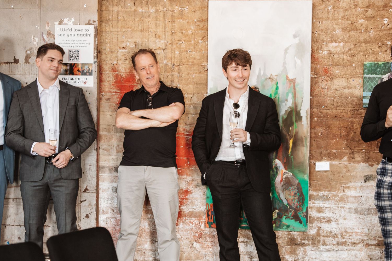 Four men standing and smiling at an art gallery event with abstract paintings on brick wall background.