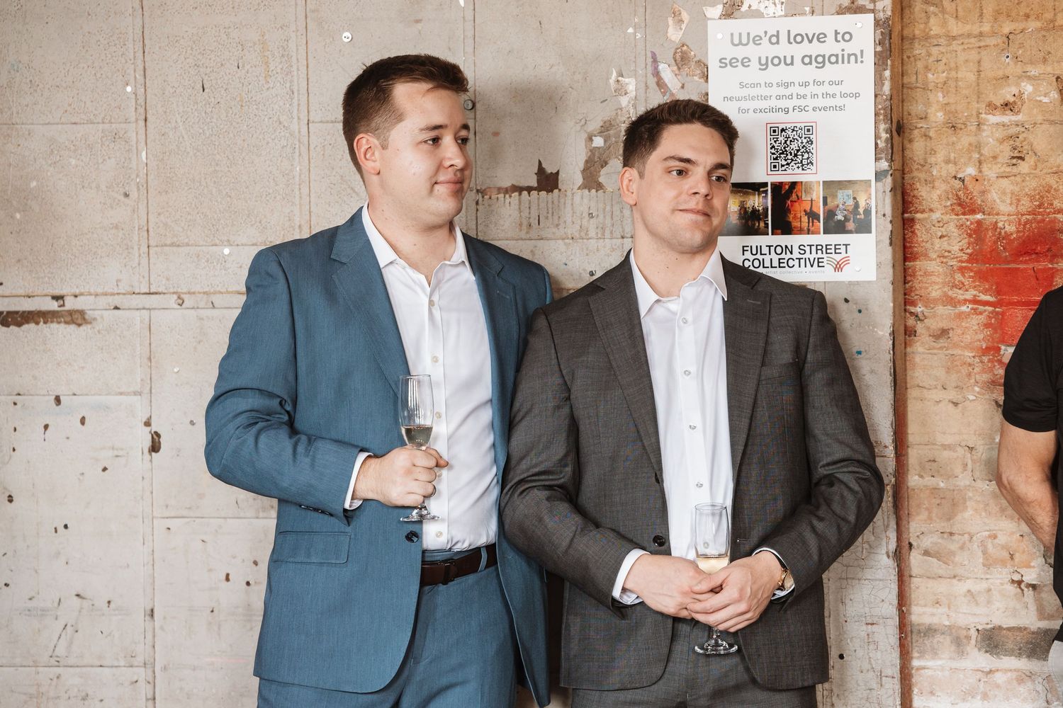Two young men in suits standing together against a concrete wall at an event.