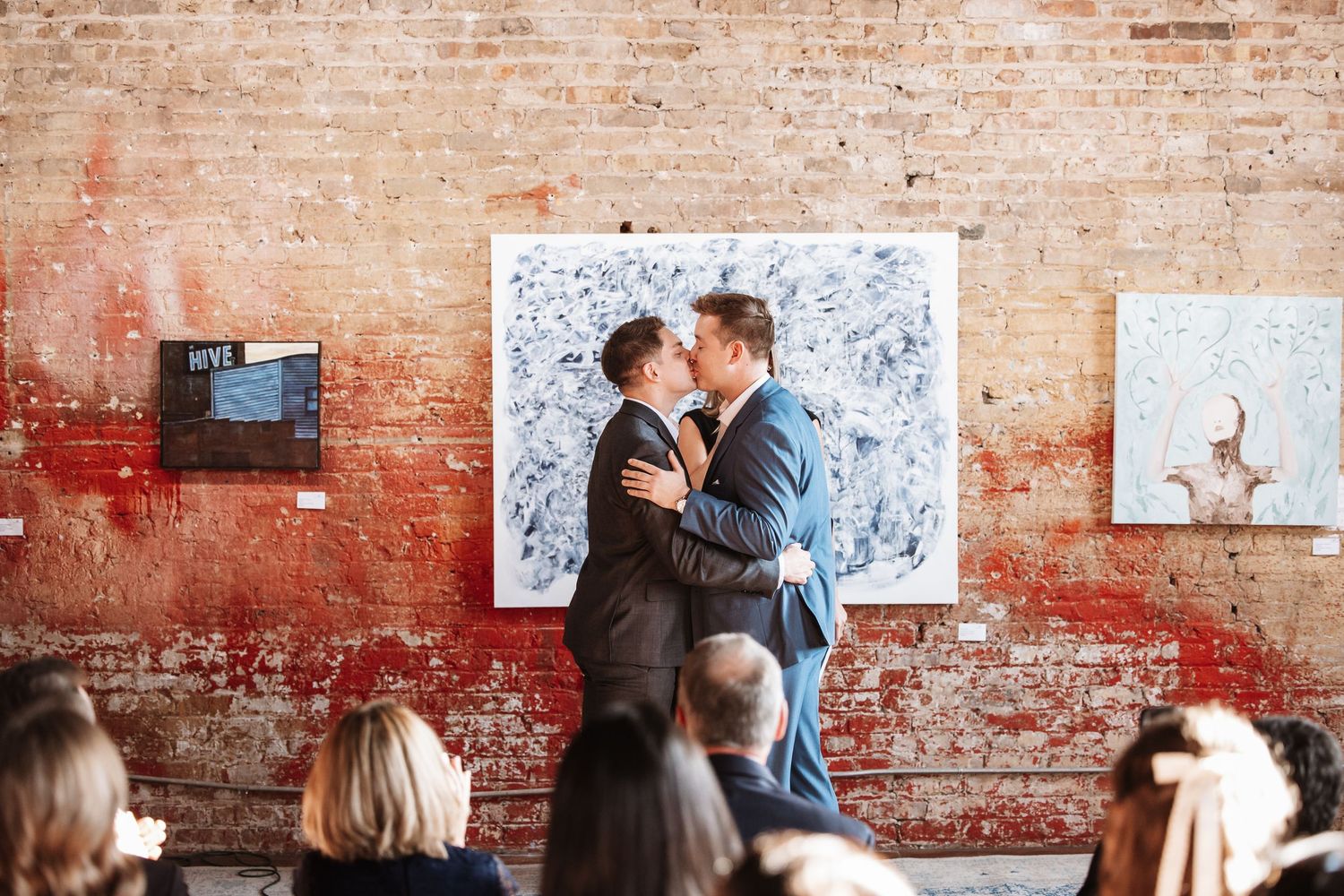 Two grooms embrace and kiss during their wedding ceremony in a rustic art gallery with brick walls.