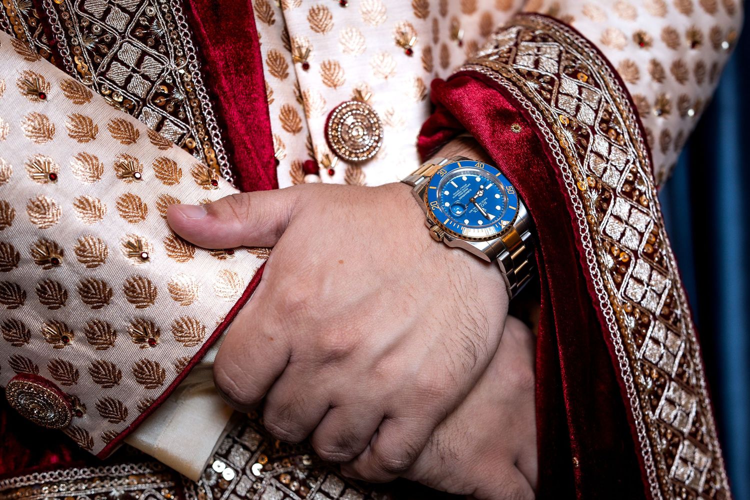 Close-up of a groom wearing a blue Rolex watch with traditional Indian wedding attire featuring gold embroidery.