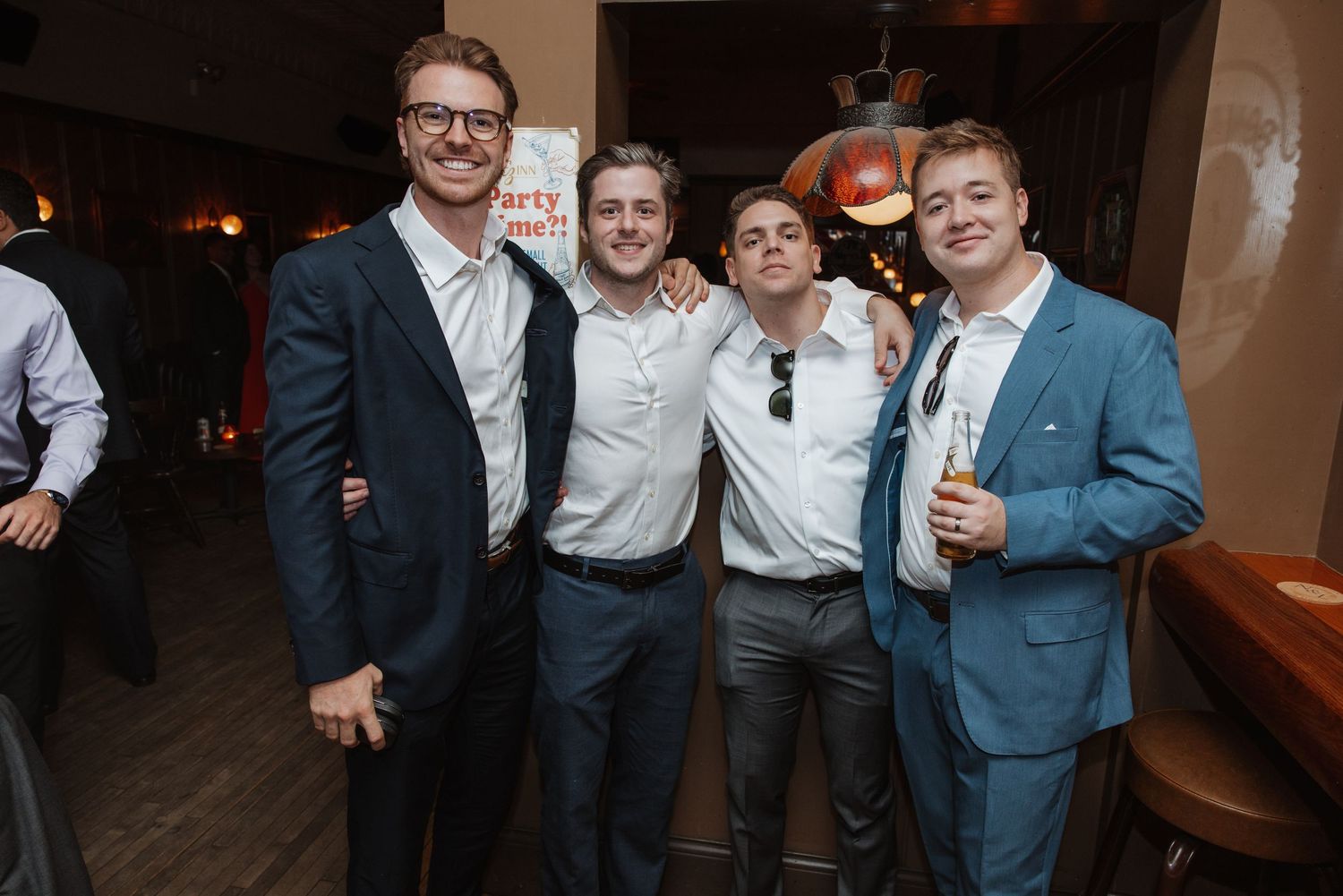 Four men in suits smiling together at an indoor wedding reception party.