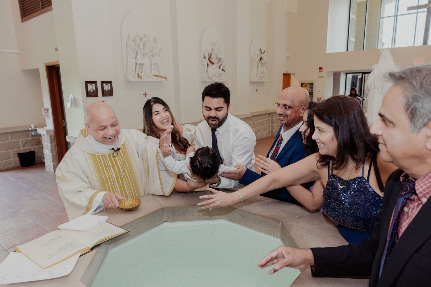 Priest performs Catholic baptism at font with family gathered around smiling baby.