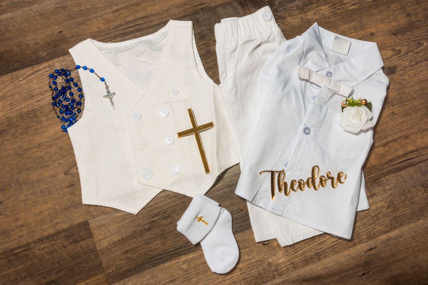 Baby baptism outfit flat lay with cross, rosary, white vest, and personalized Theodore gown.