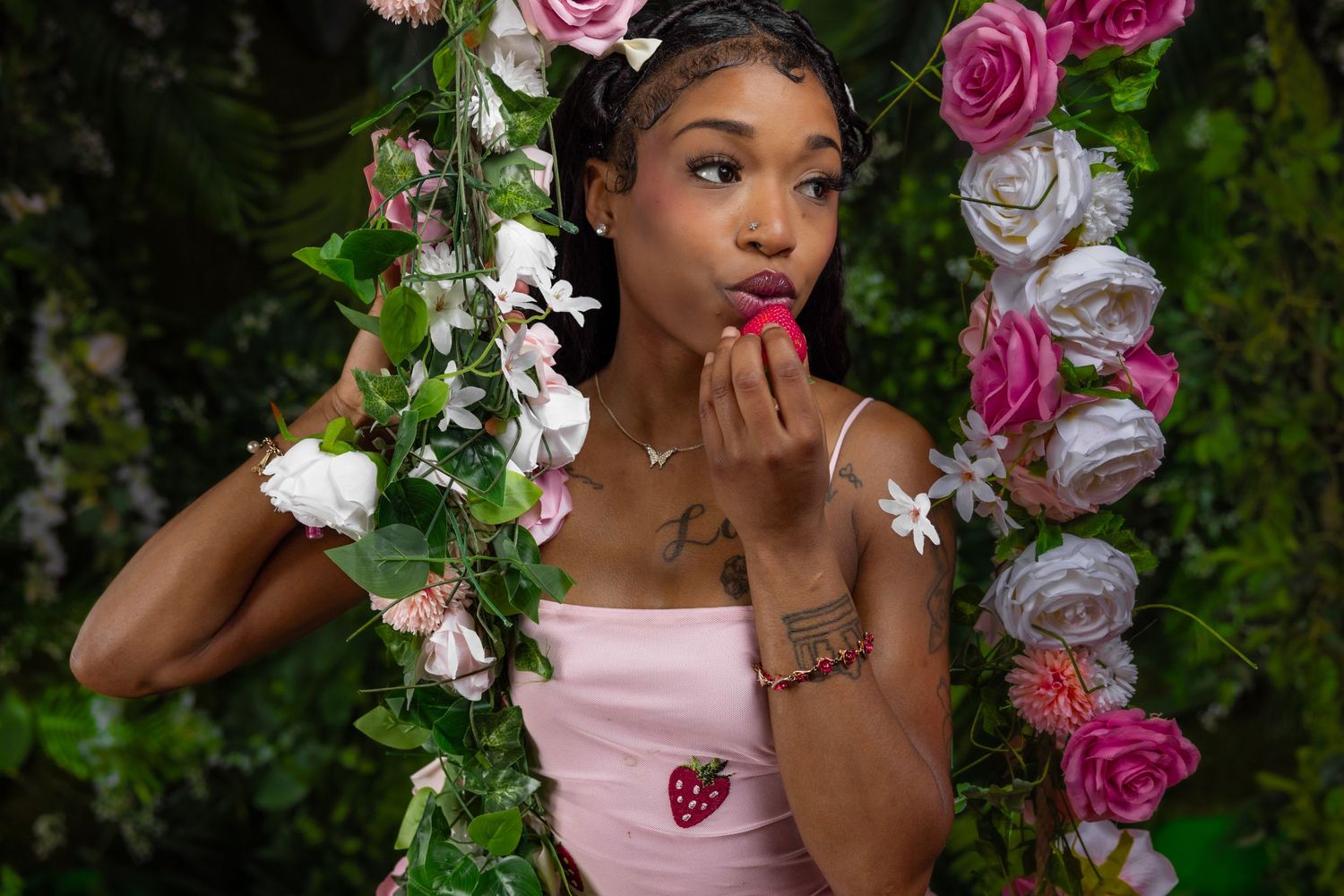 Young Black woman in pink strawberry dress holds strawberry near lips on floral swing backdrop.
