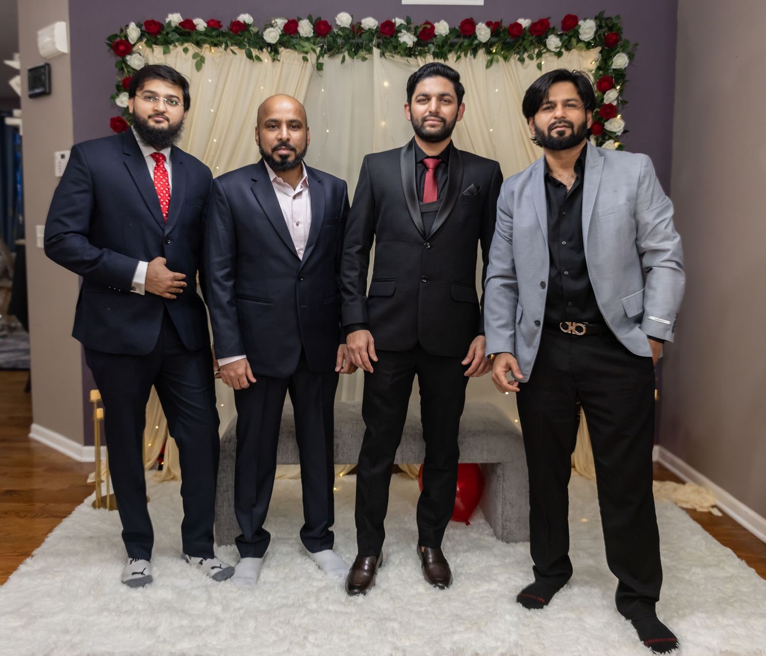 Four men in formal suits pose together at an elegant wedding venue decorated with red and white flowers.