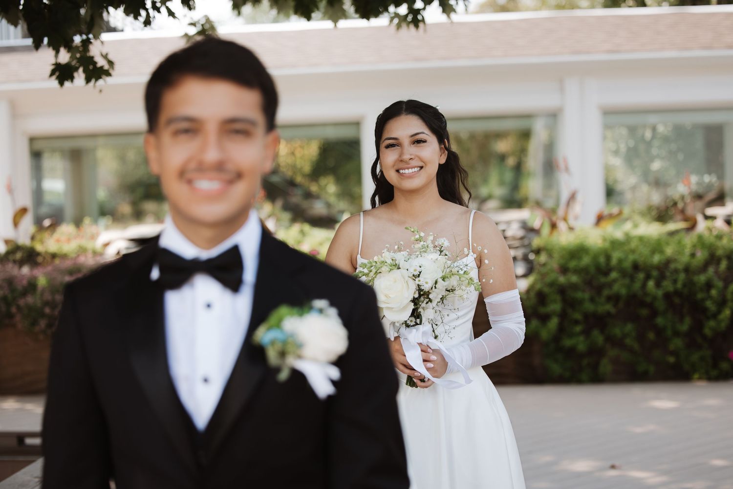 Smiling bride in white dress with bouquet stands behind groom in black tuxedo outdoors.