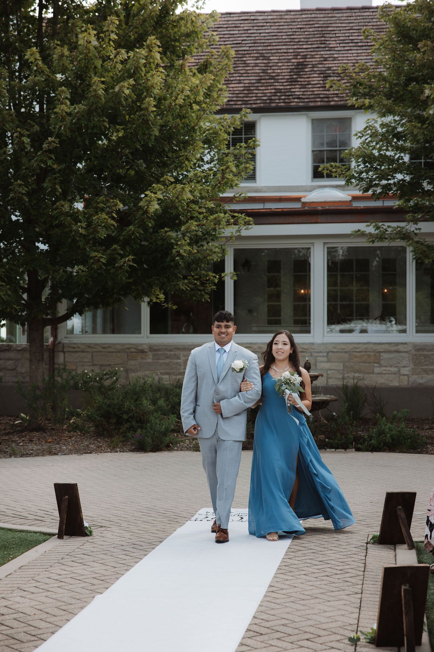 Groomsman escorts bridesmaid in flowing blue dress along white aisle runner at outdoor wedding.