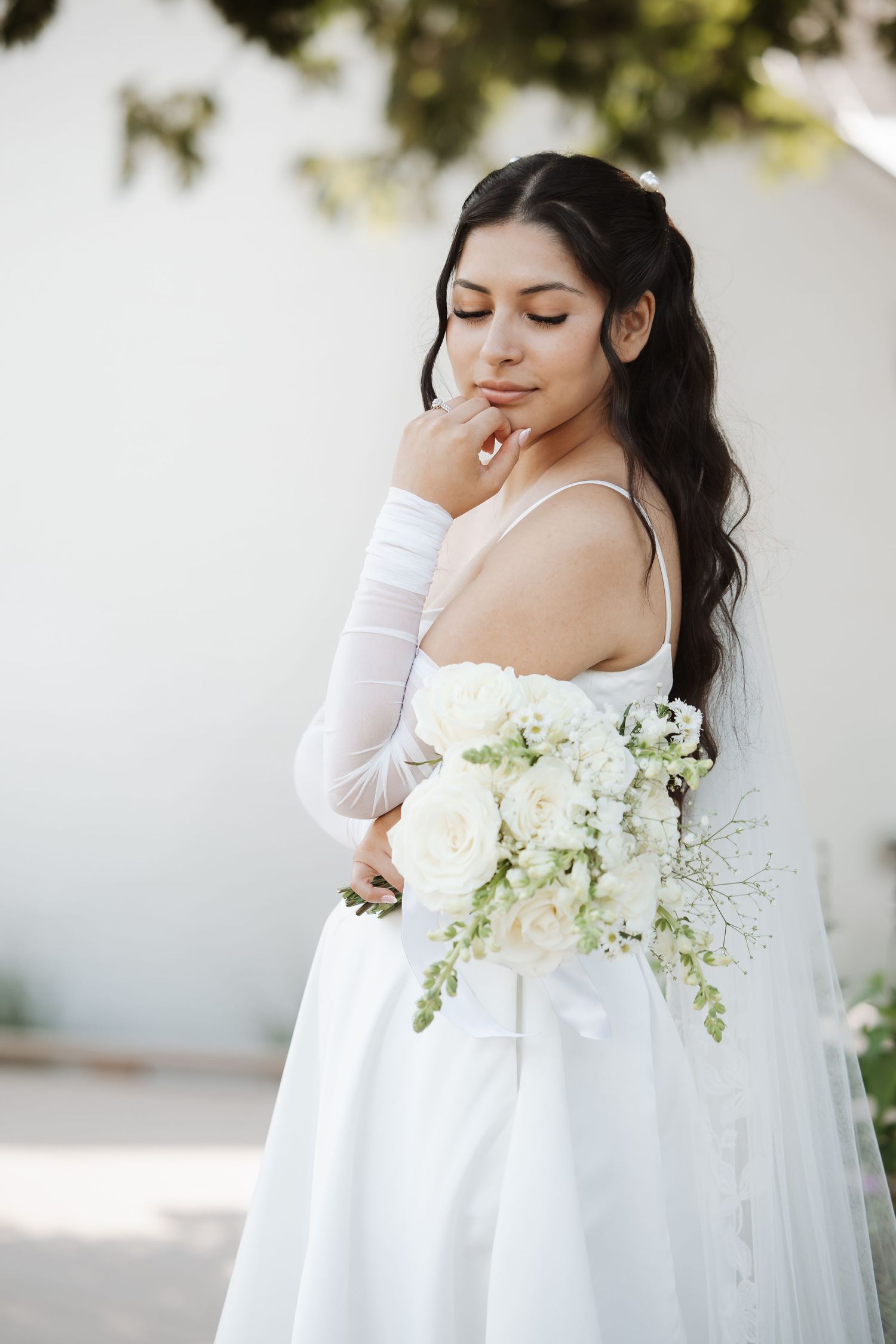 Elegant bride in white off-shoulder gown with gloves holds white rose bouquet, eyes downcast outdoors.
