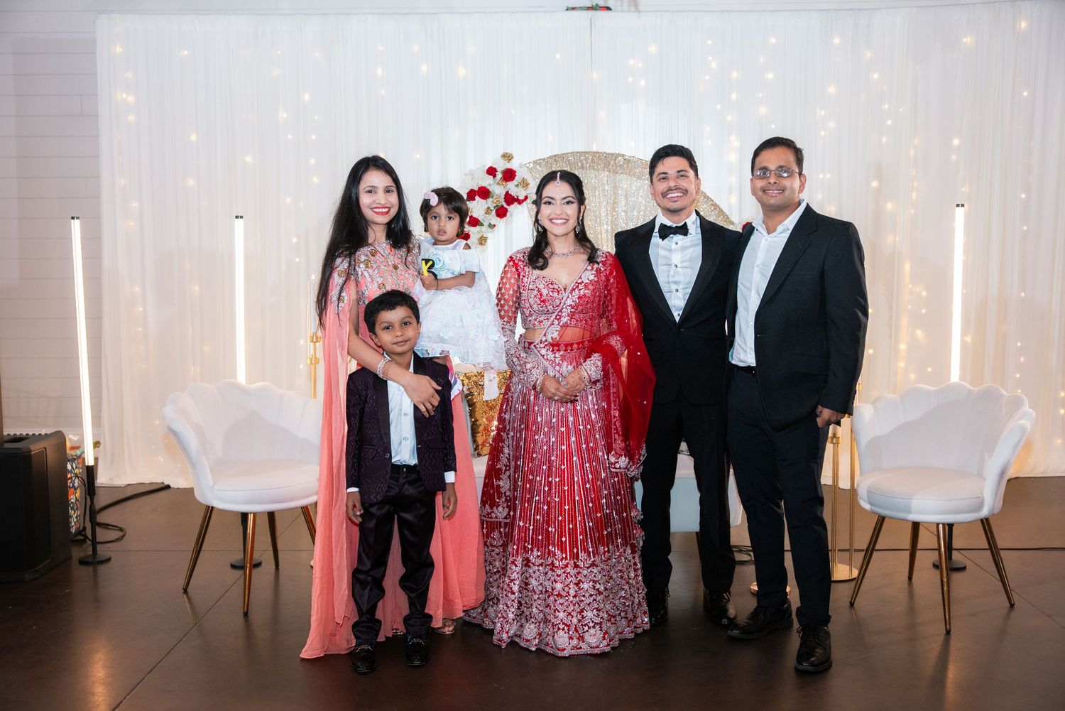 Family of five posing with bride in red bridal lehenga and groom in tuxedo at wedding venue.