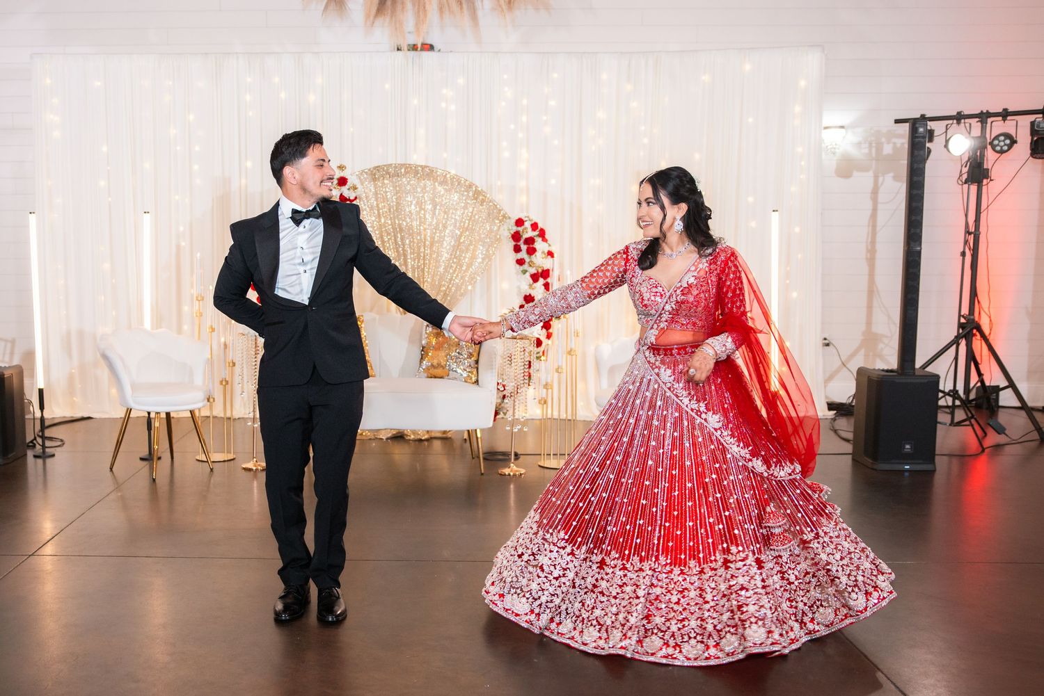 Groom in black tuxedo holds hand of bride in red lehenga as she twirls at Indian wedding reception.