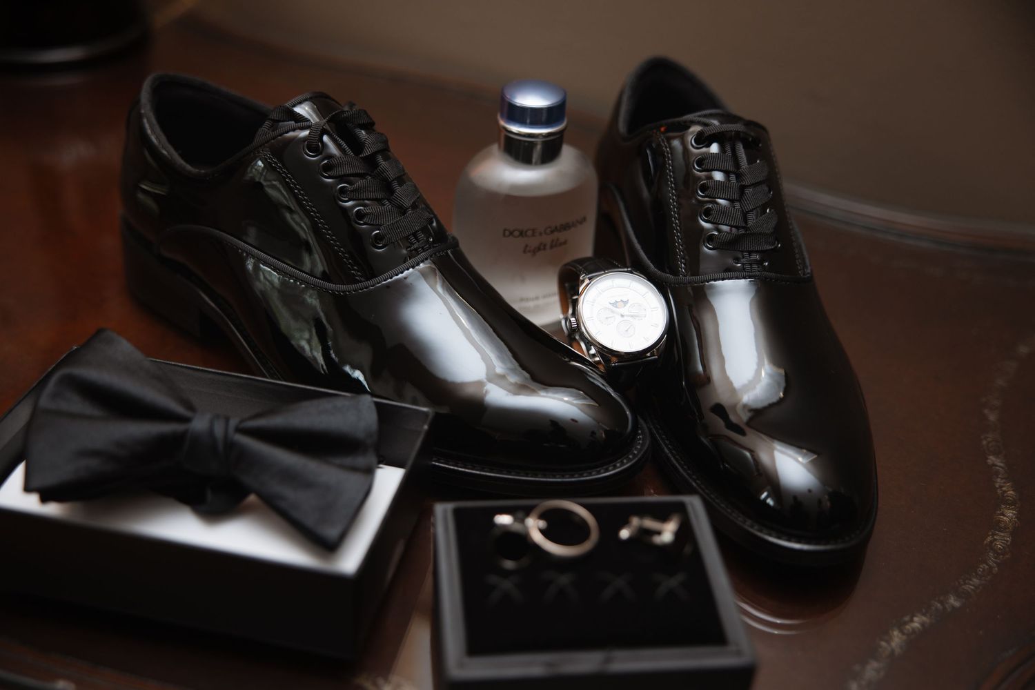 Groom's black patent shoes, bow tie, watch, and cufflinks styled on table.