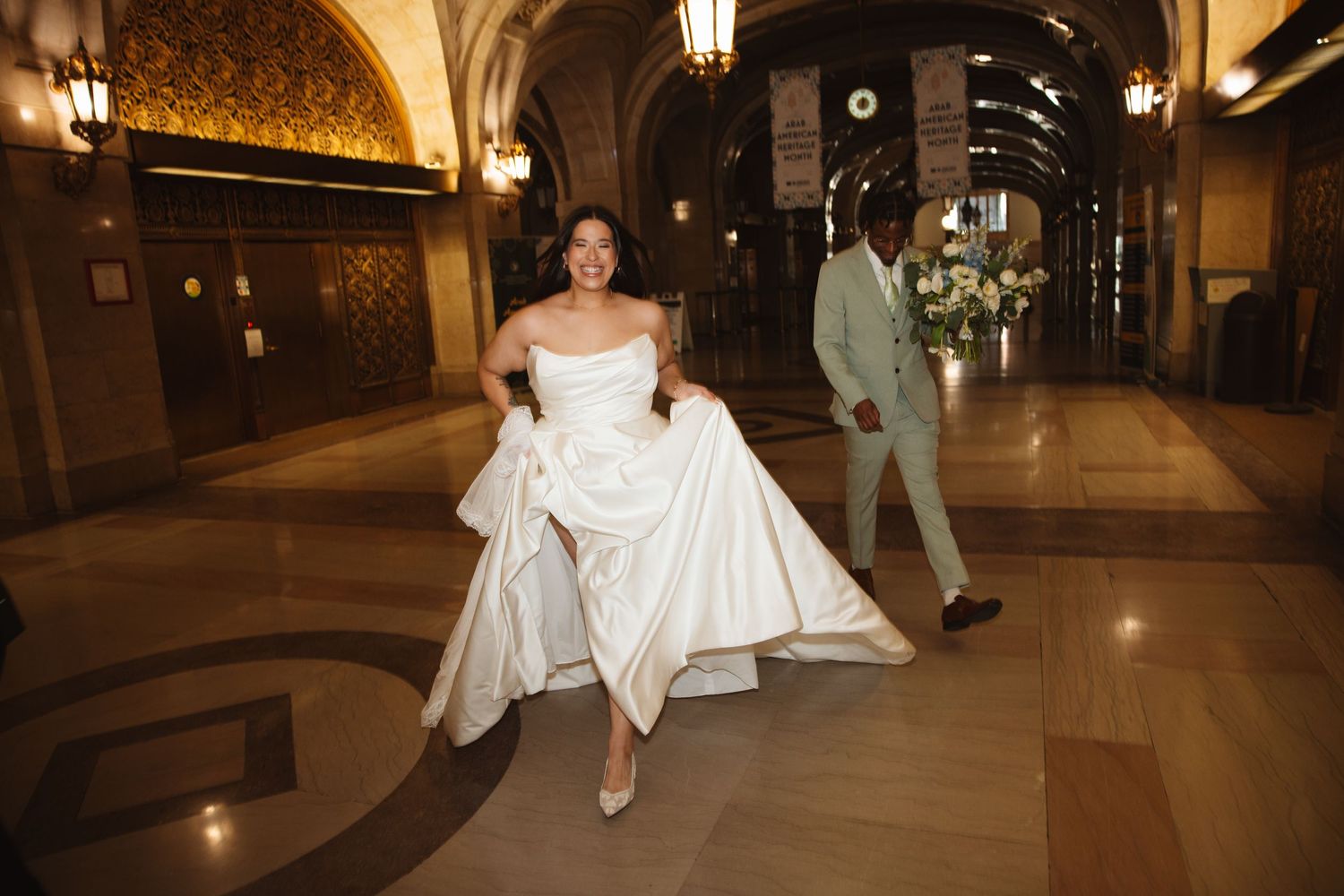 Bride lifts white wedding gown while walking through ornate hotel corridor with groom following behind.