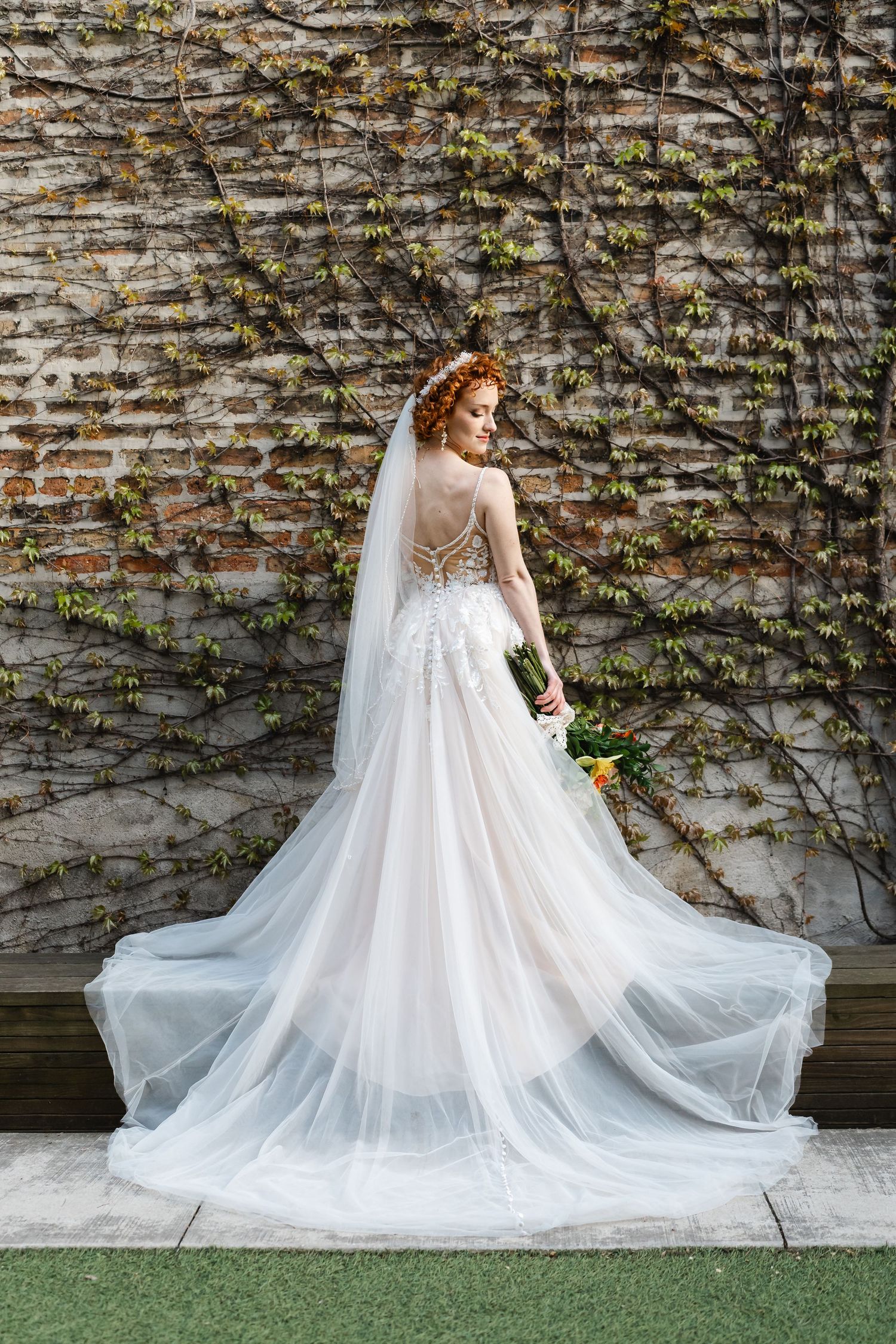Red-haired bride in lace-back white tulle wedding gown with cathedral train stands before ivy-covered brick wall.