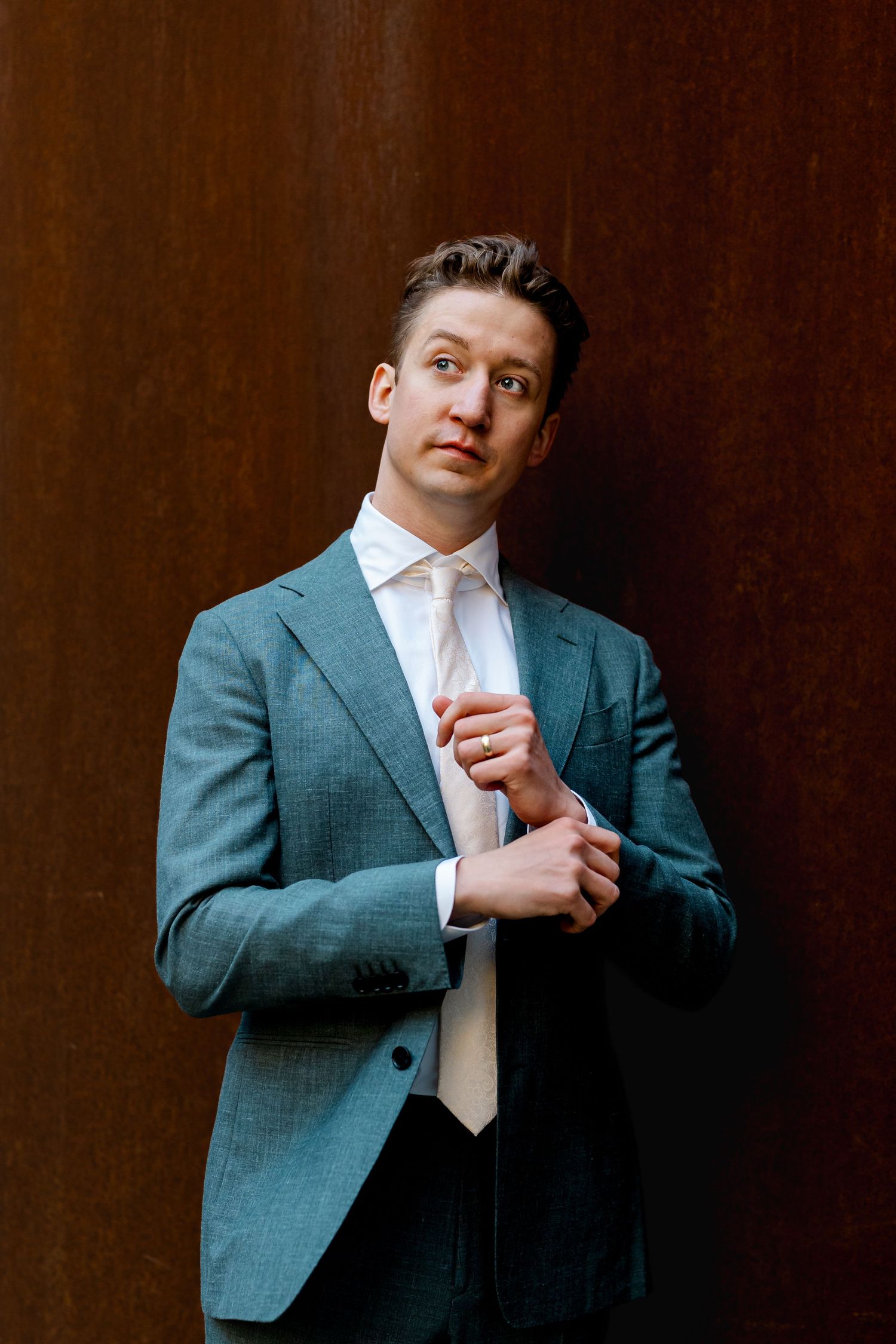 Groom in grey suit adjusts cufflinks while posing against rustic brown metal wall before wedding.