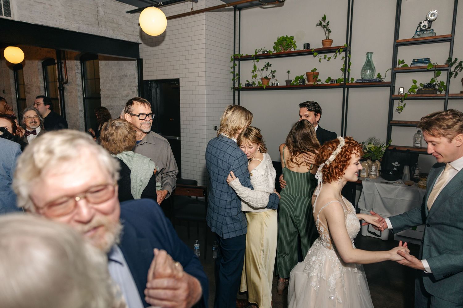 Guests dance and mingle at lively indoor wedding reception with bride visible in background.