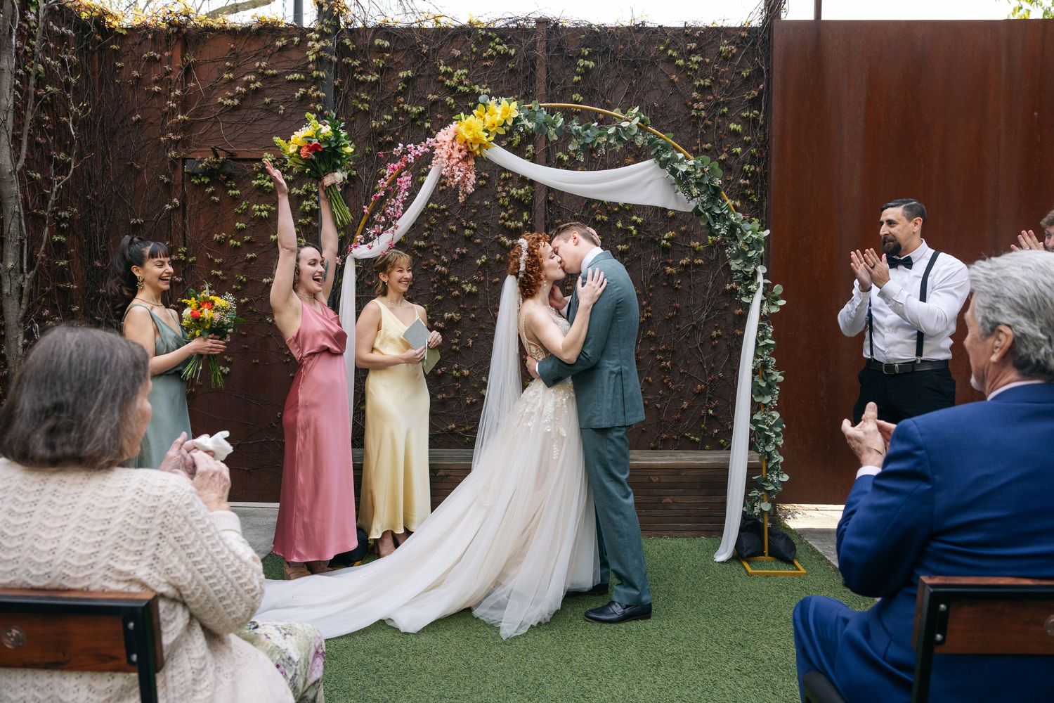 Bride and groom share first kiss at outdoor wedding ceremony under floral arch with cheering wedding party.