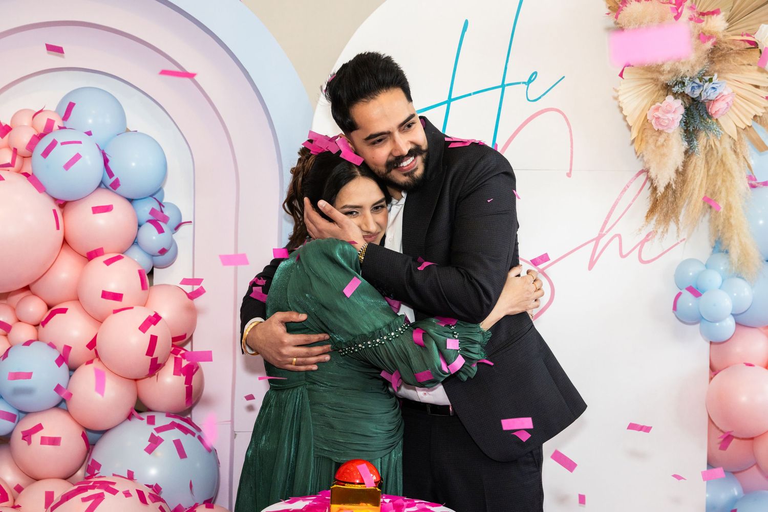 Emotional couple hugging at gender reveal party with pink confetti and balloon backdrop.