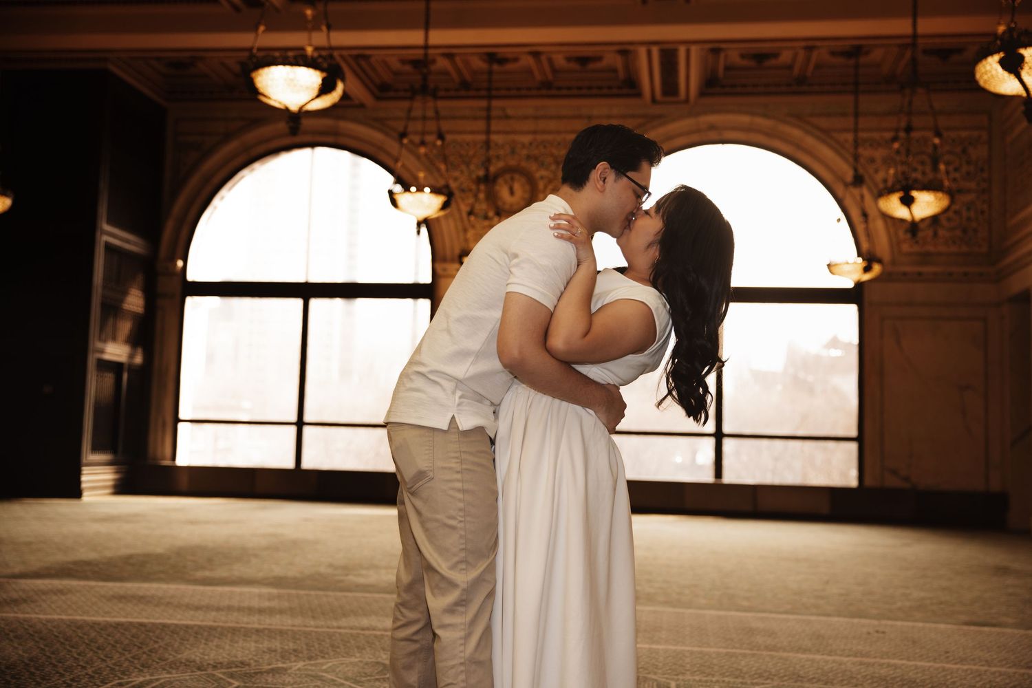 Couple shares romantic kiss in sunlit ballroom with arched windows.