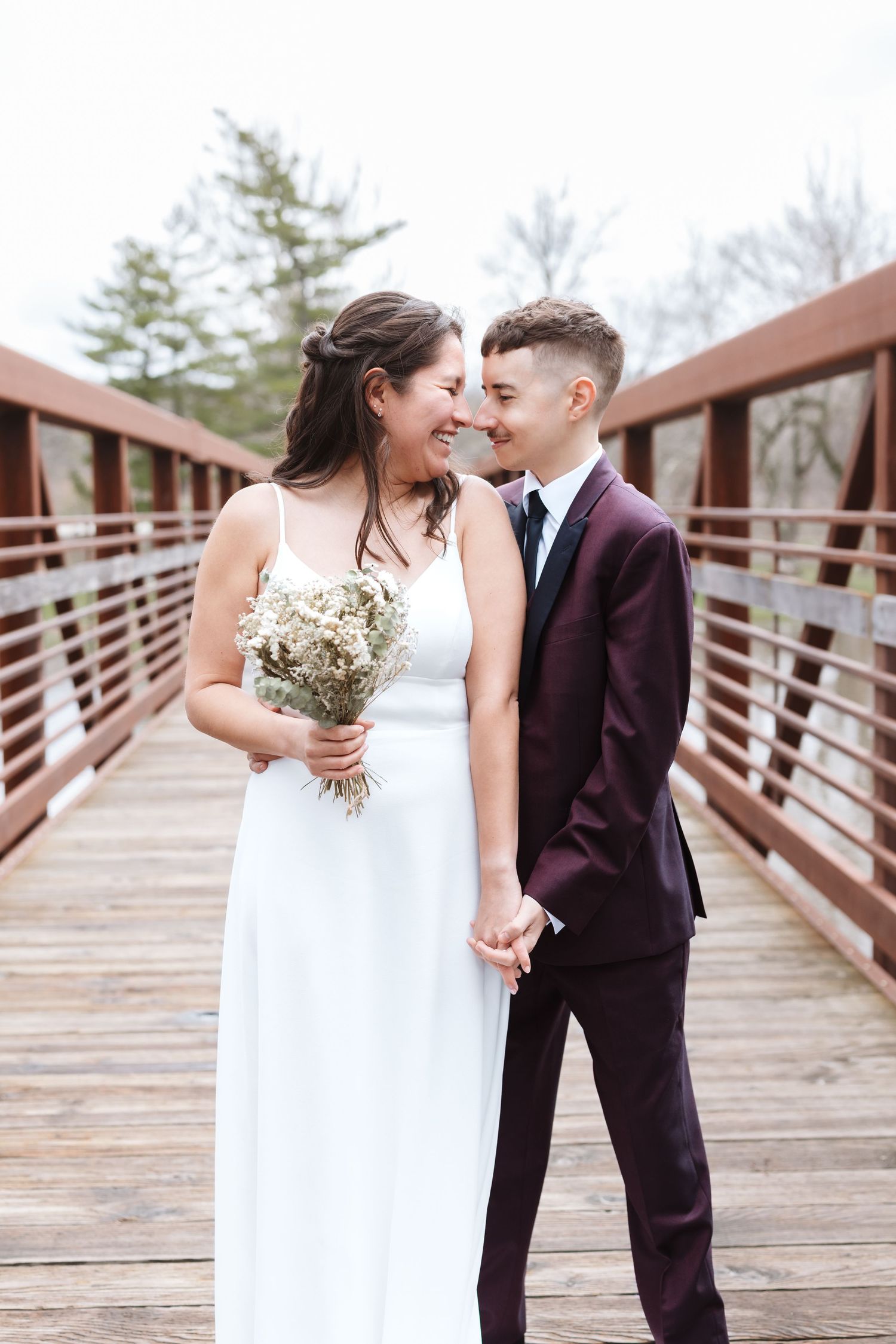 A bride in a white gown holds a bouquet while sharing a romantic moment with their partner on a wooden bridge.
