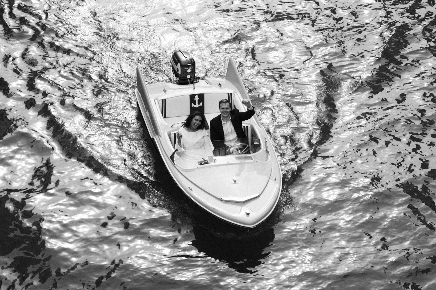 Aerial black and white photo of a small speedboat cutting through rippling water with two passengers aboard.