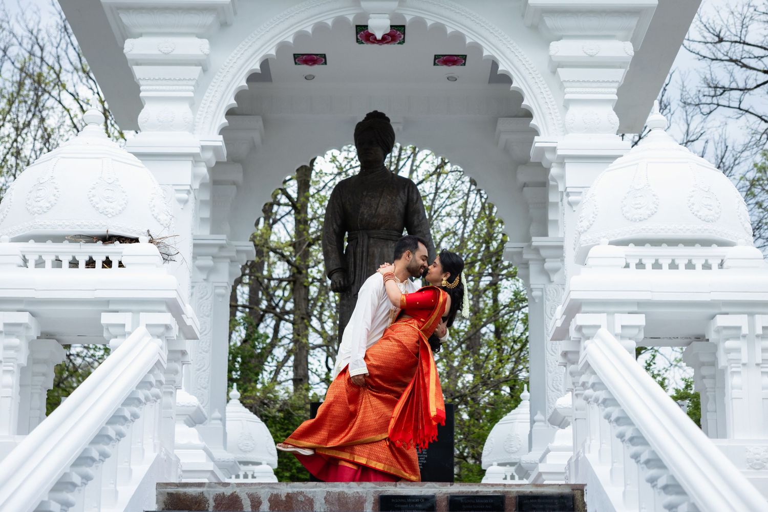 Couple sharing a romantic dip kiss in front of a white ornate Hindu temple archway with a bronze statue.