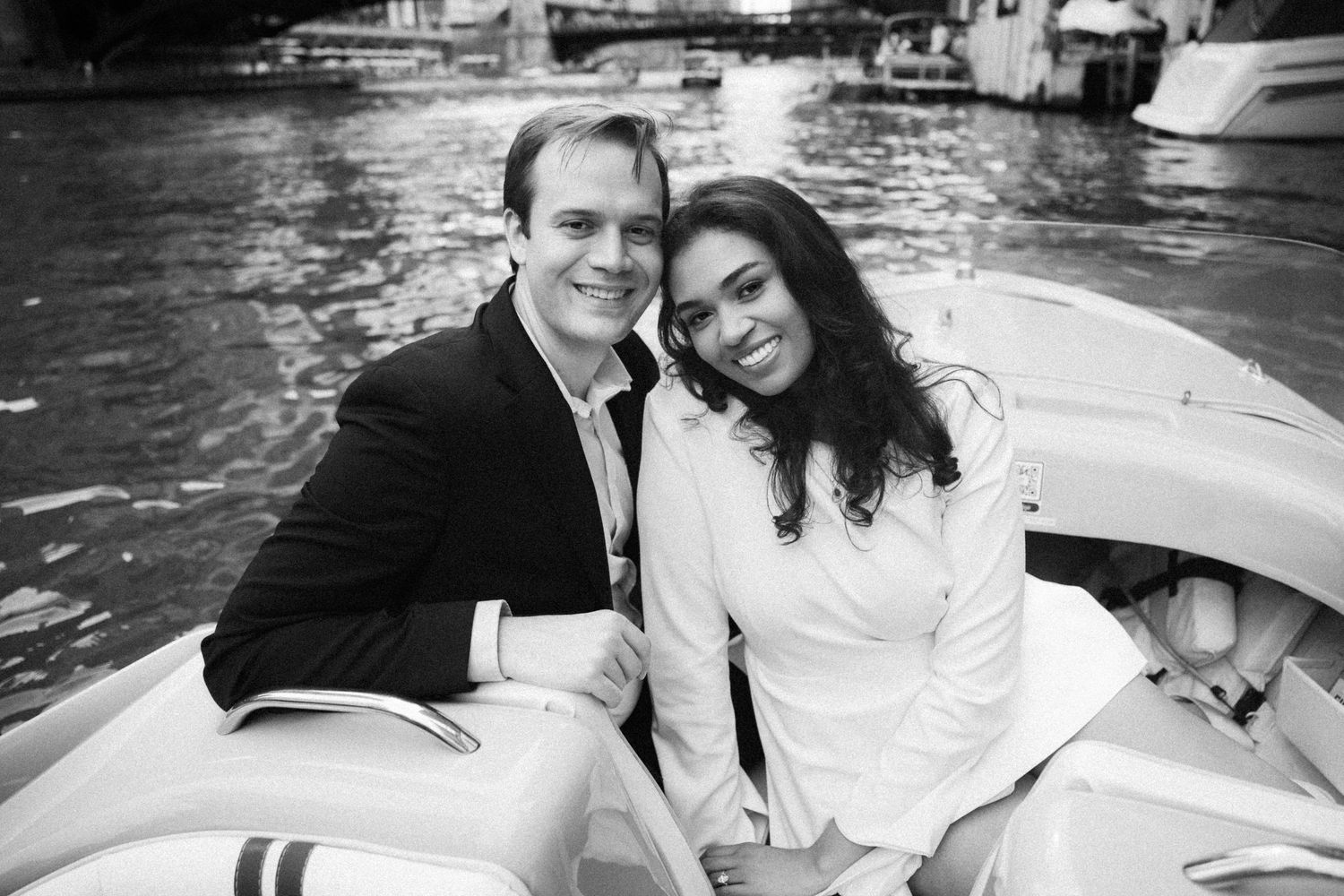 A smiling couple poses together on a boat on a river in a black and white engagement photo.