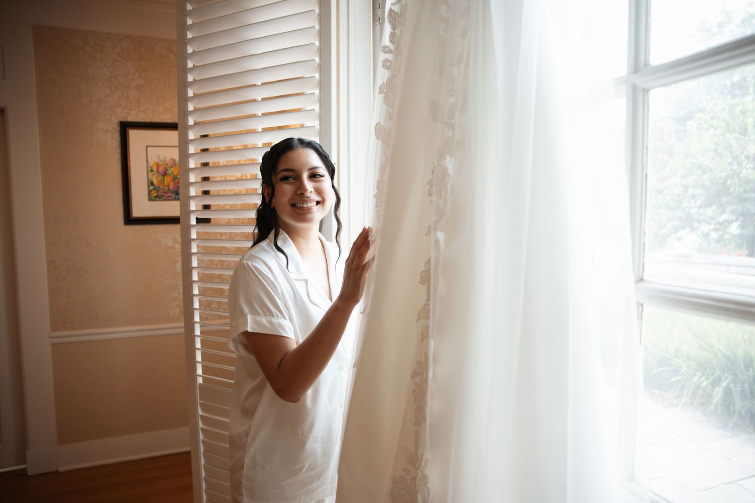 Smiling dark-haired bride in white robe touching her lace wedding gown hanging near a sunlit window.
