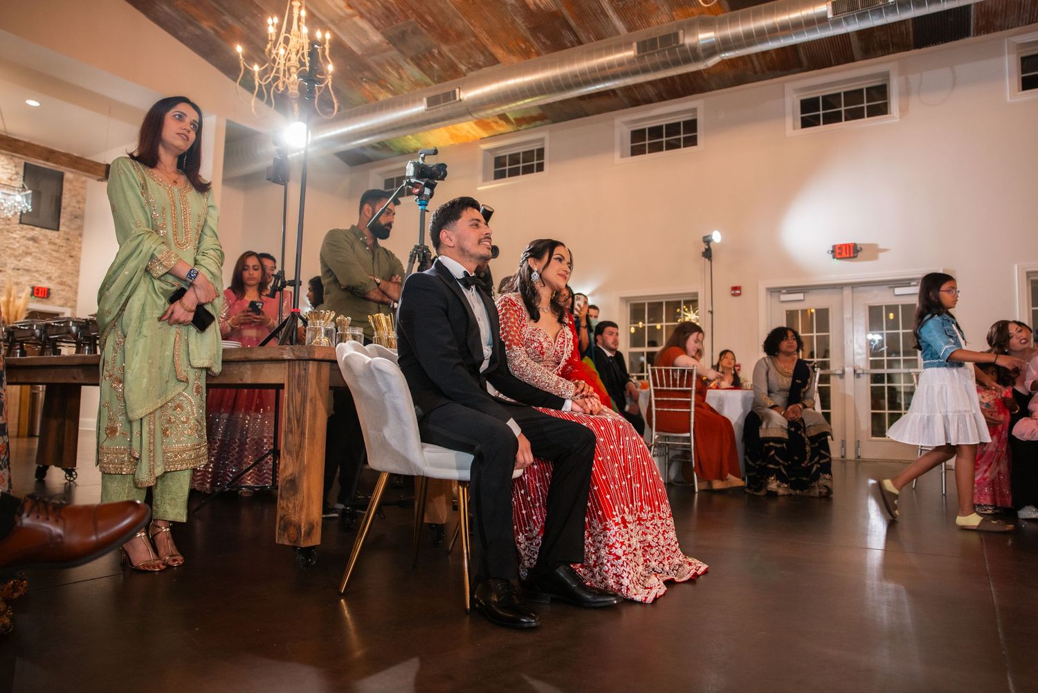 Bride in red lehenga and groom in tuxedo seated at South Asian wedding reception.