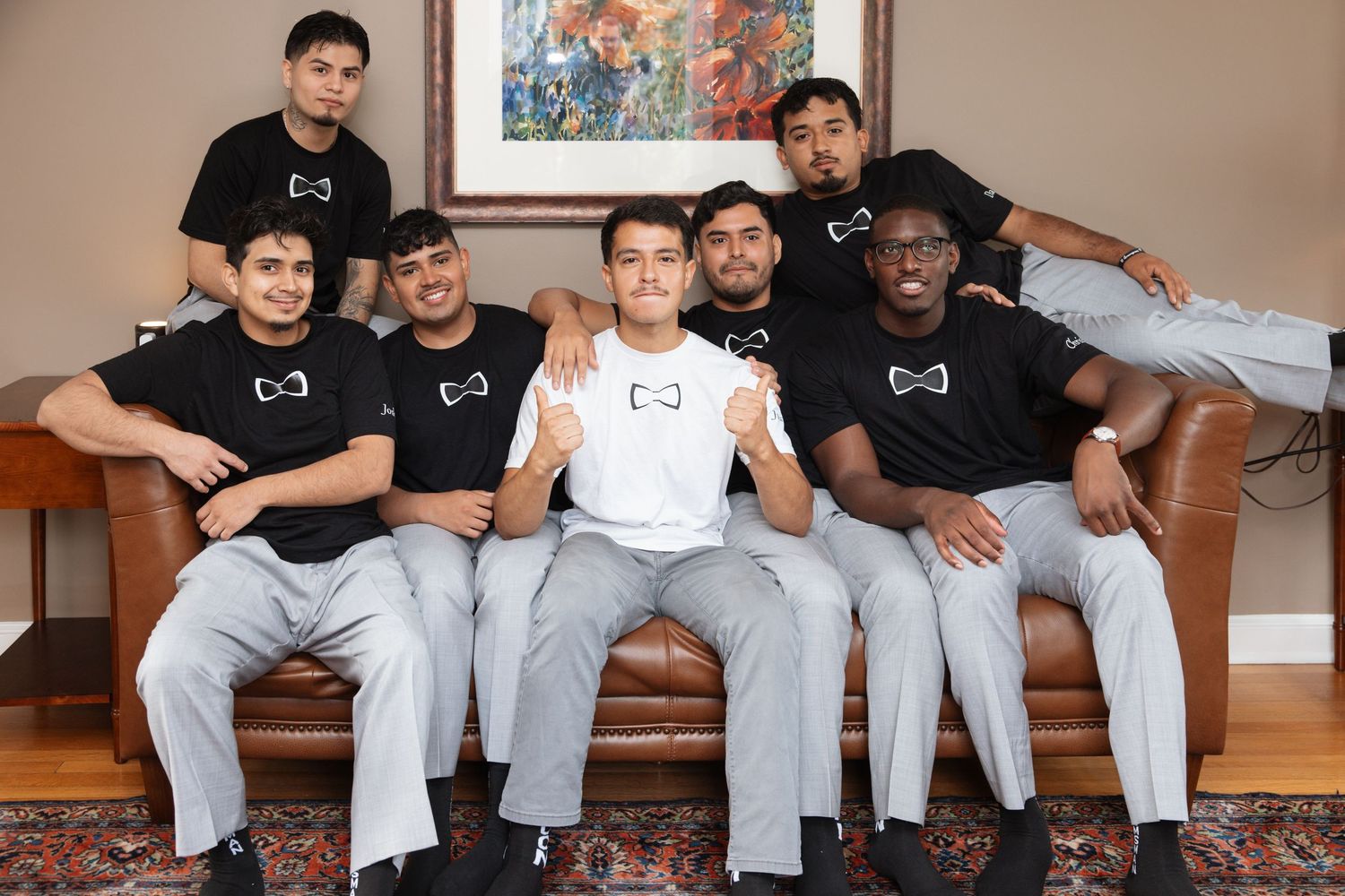Seven young men in matching black and white logo t-shirts and gray pants pose together on a brown sofa.