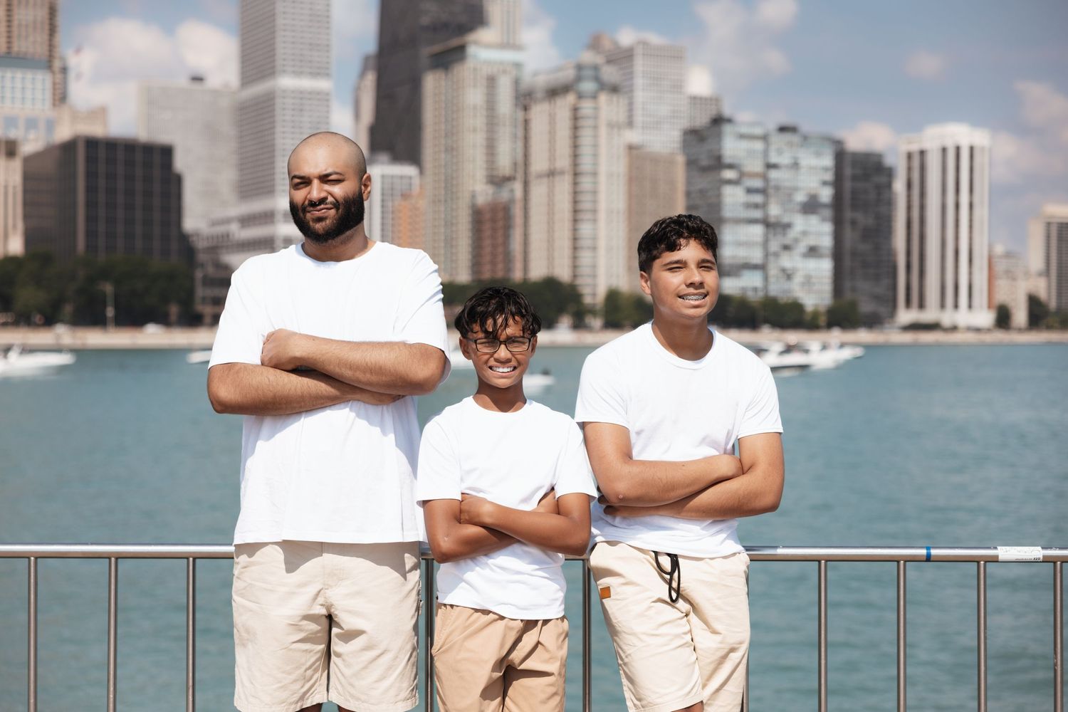Three males in matching white shirts stand arms crossed by Chicago lakefront skyline on clear day.