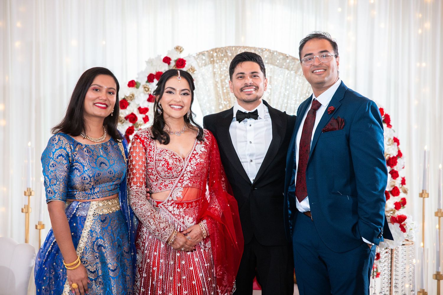 Indian wedding group photo with bride in red lehenga, groom in tuxedo, and two family members in ethnic formal wear.