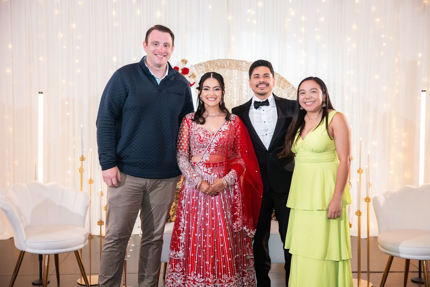Bride in red lehenga and groom in tuxedo pose with two guests at a romantic wedding reception.