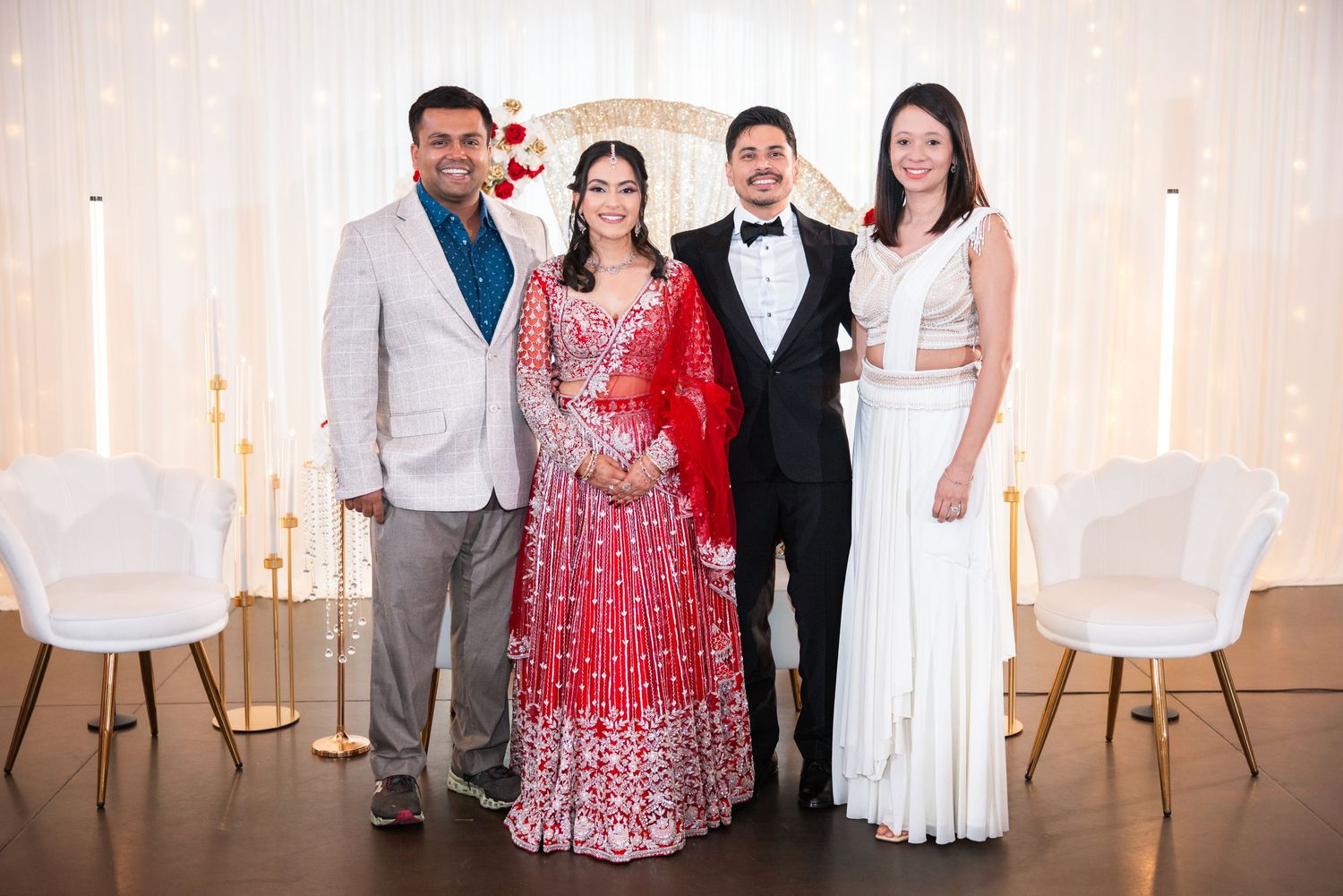 Couple in traditional wedding attire poses with two smiling guests at a fairy-lit reception.