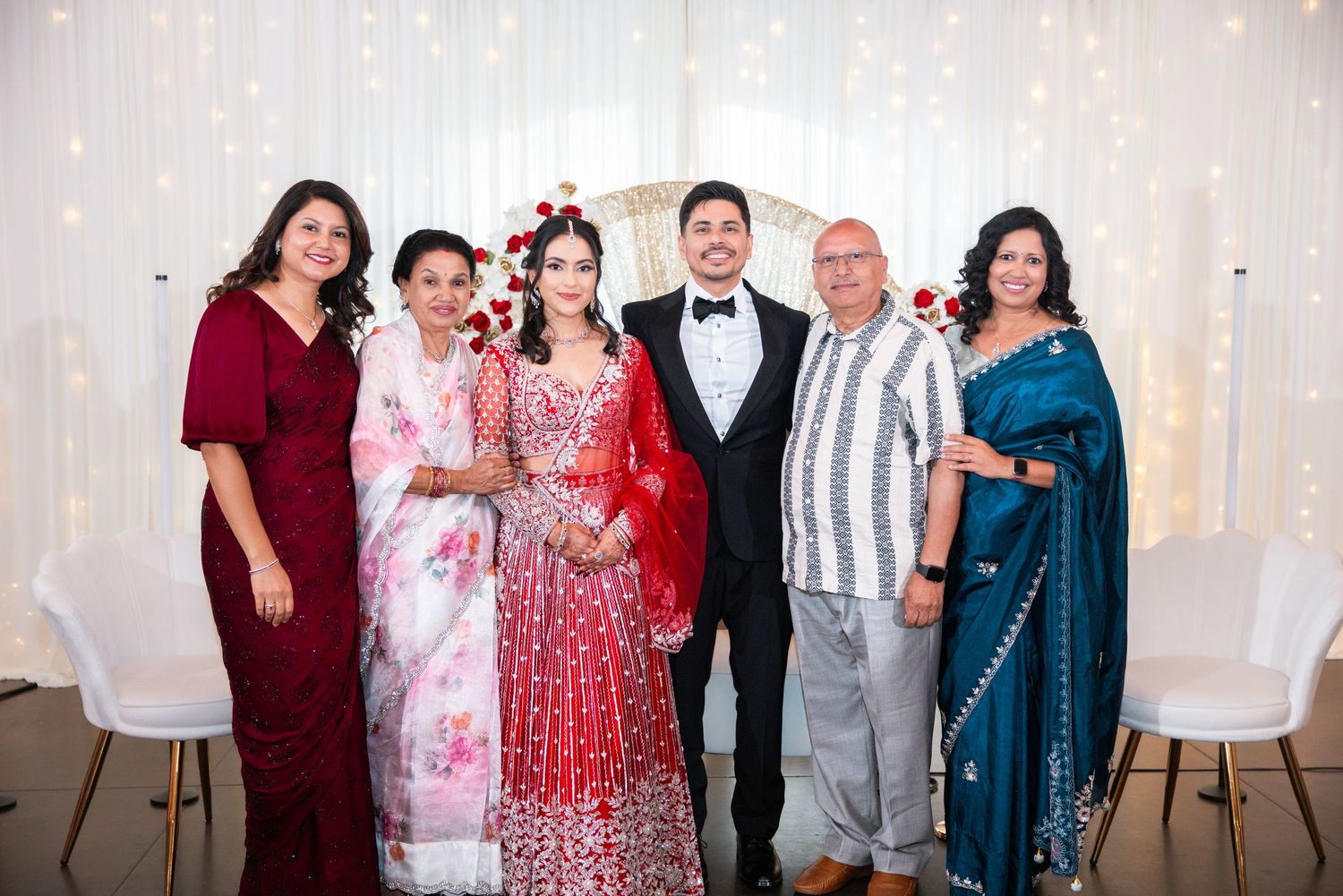 South Asian wedding group photo with bride in red lehenga, groom in tuxedo, and four family members posing together.