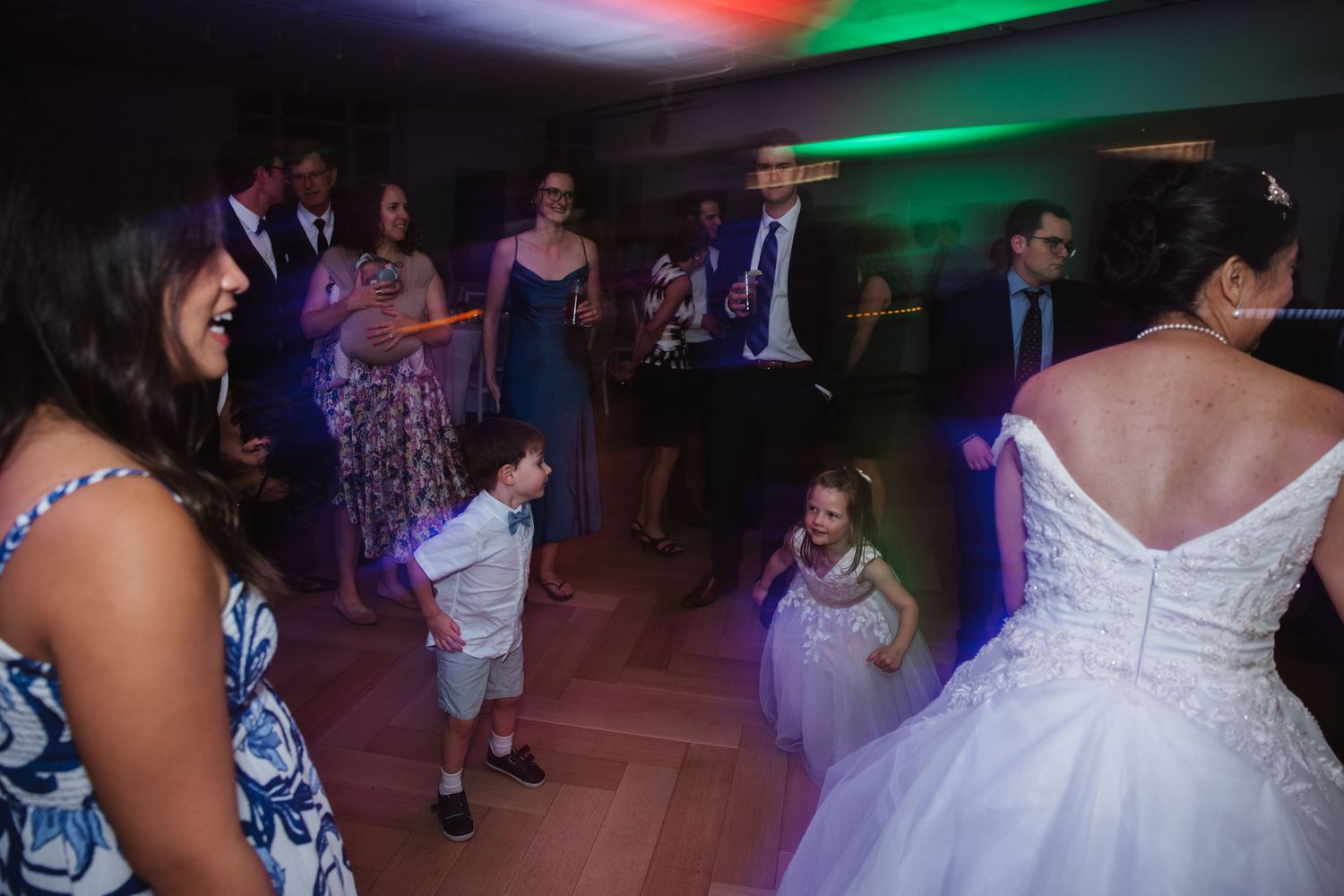 A bride in a white gown dances with children and guests on a vibrant, colorfully lit wedding dance floor.