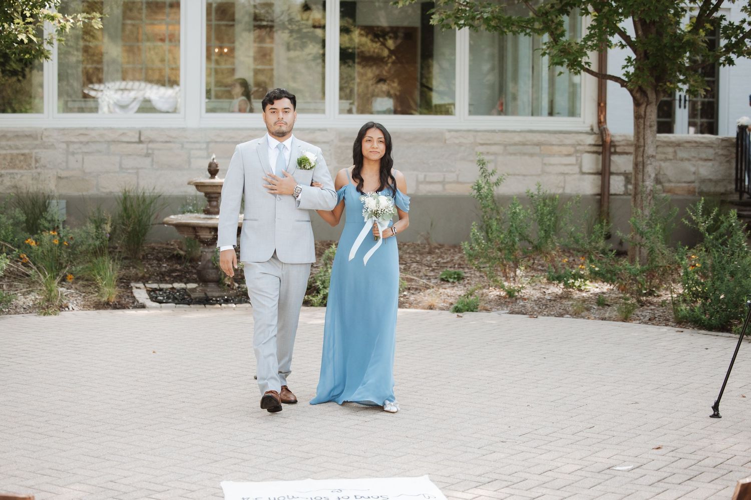 Groomsman in gray suit escorts bridesmaid in blue gown down outdoor wedding ceremony aisle.