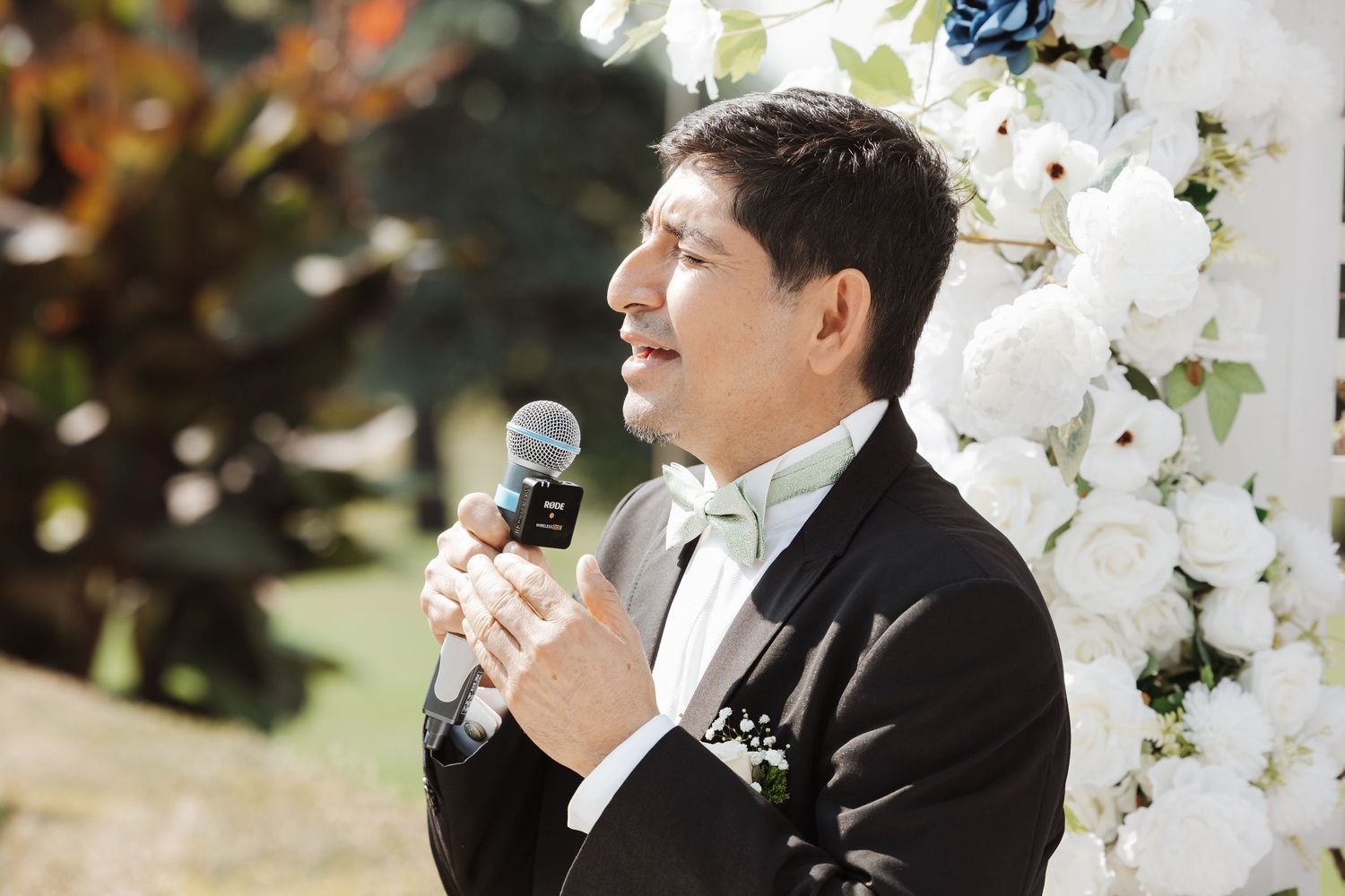 Officiant in black tuxedo speaks into microphone at outdoor wedding ceremony near floral arch.