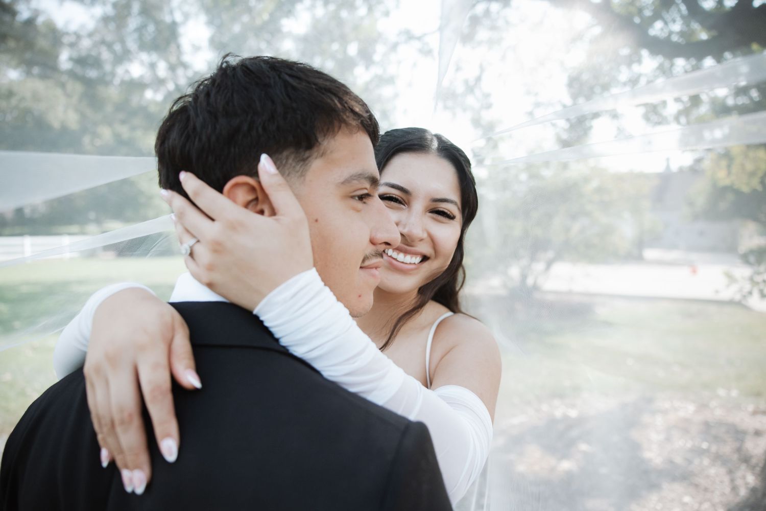 Happy bride embracing groom outdoors, smiling warmly in white wedding dress on sunny day.