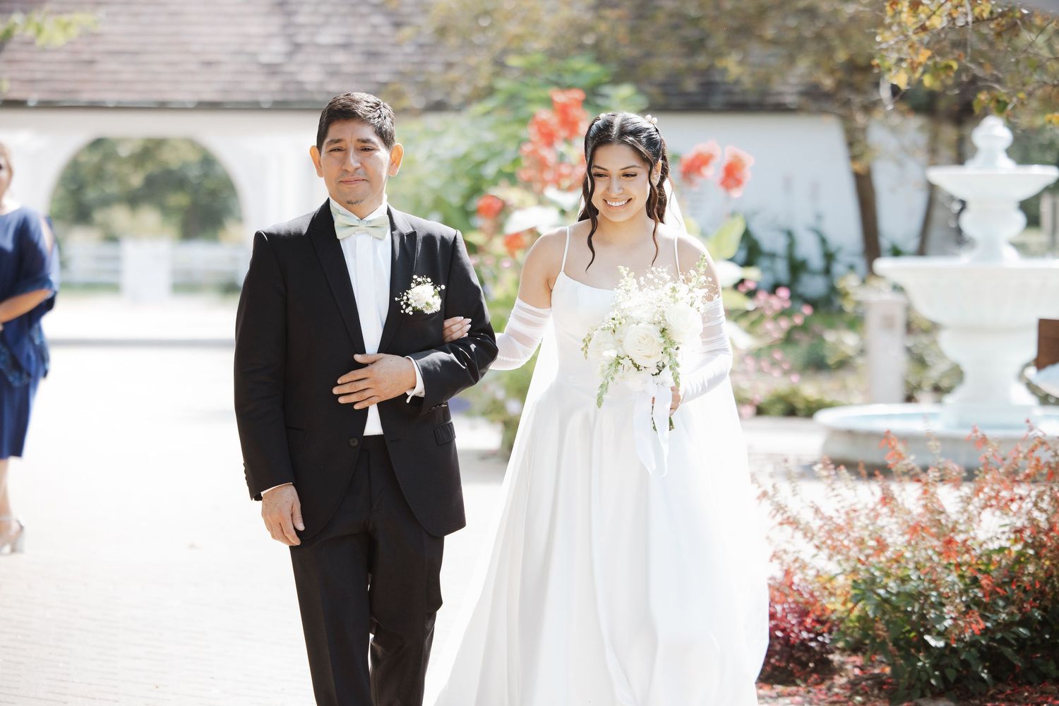 Smiling bride in white gown walks arm-in-arm with escort at sunny outdoor wedding ceremony.