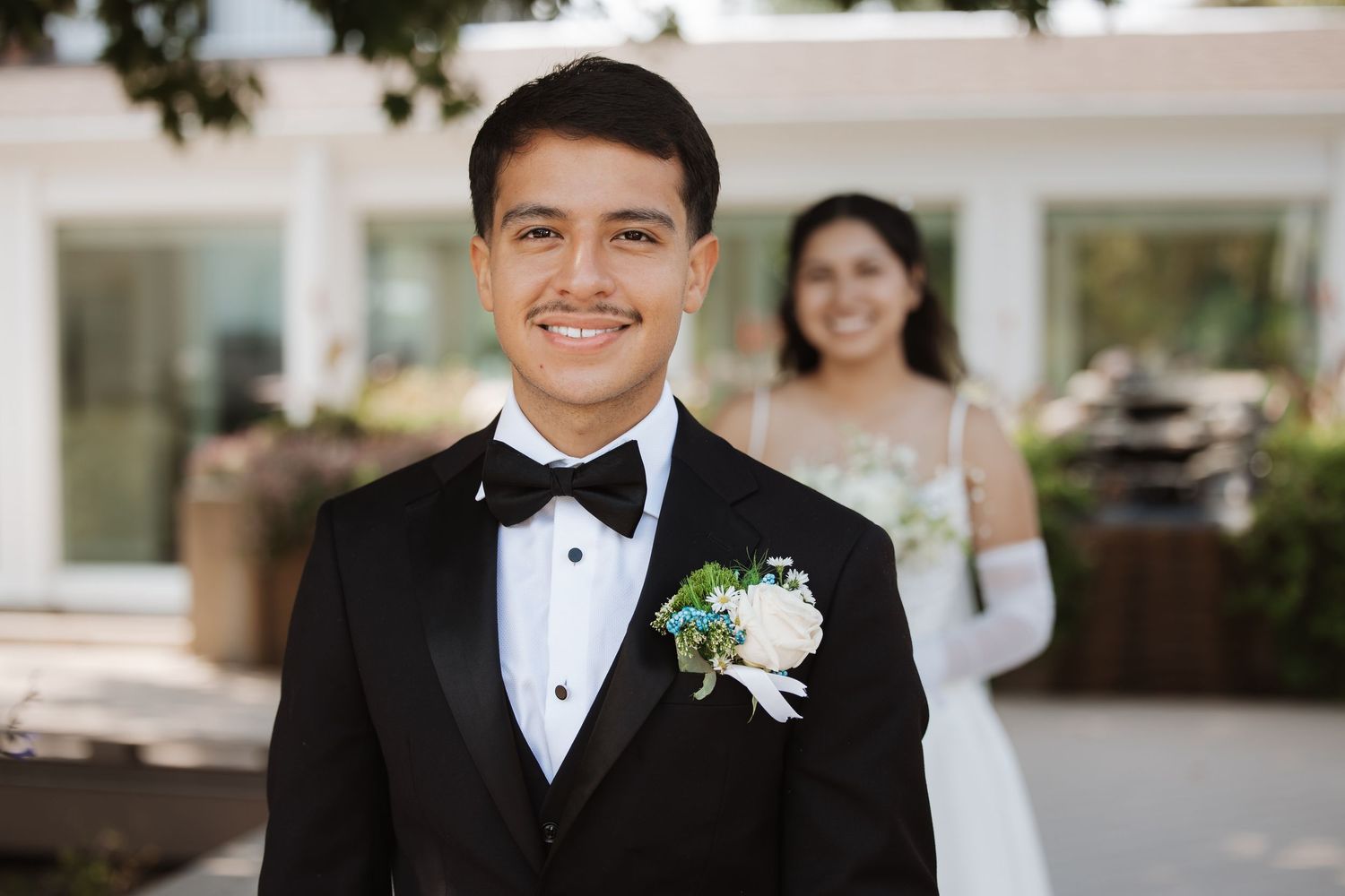 Groom in black tuxedo smiles with bride in white dress visible in background outdoors.