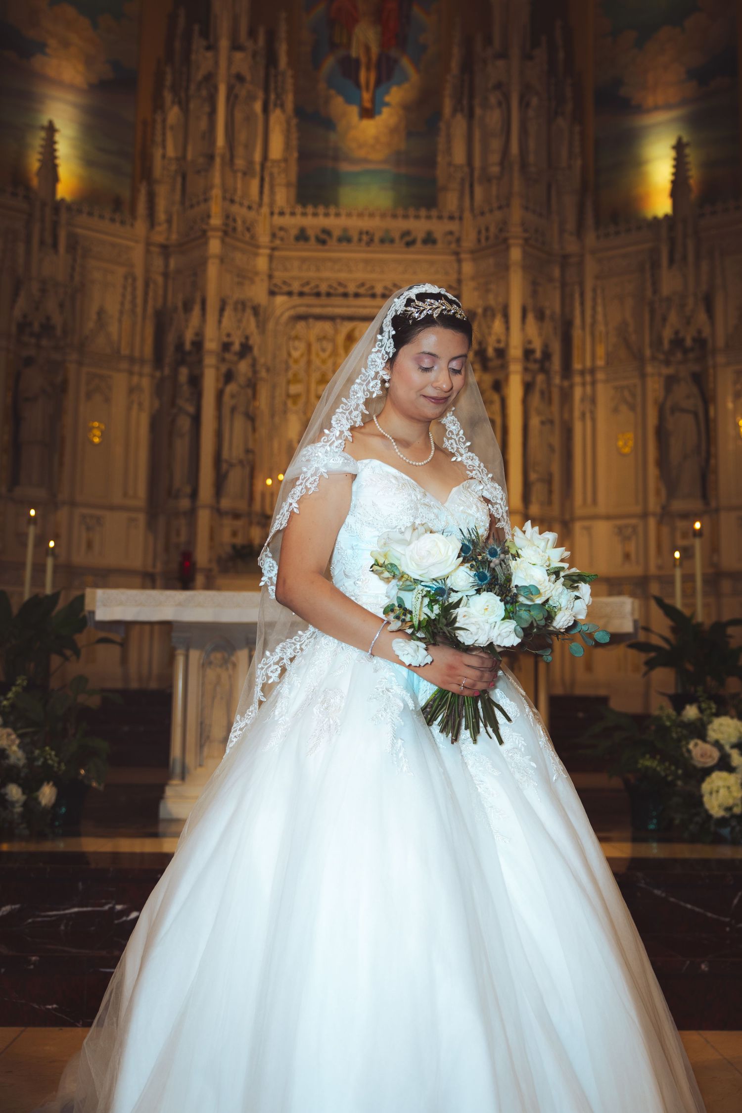 Bride in white lace ball gown and veil holding white bouquet at ornate cathedral altar.