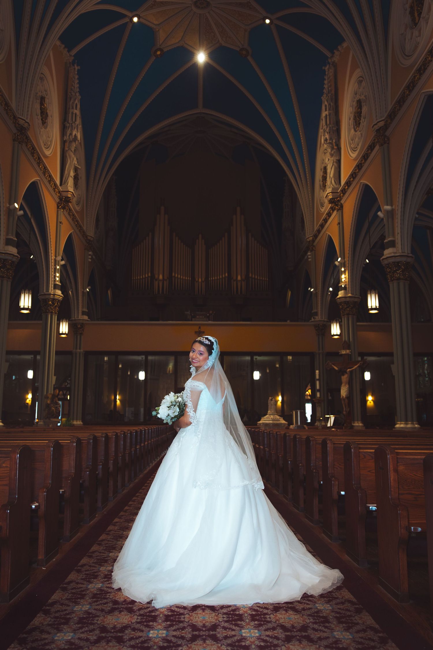 Bride in white ball gown stands in grand church aisle with vaulted arches and pipe organ backdrop.